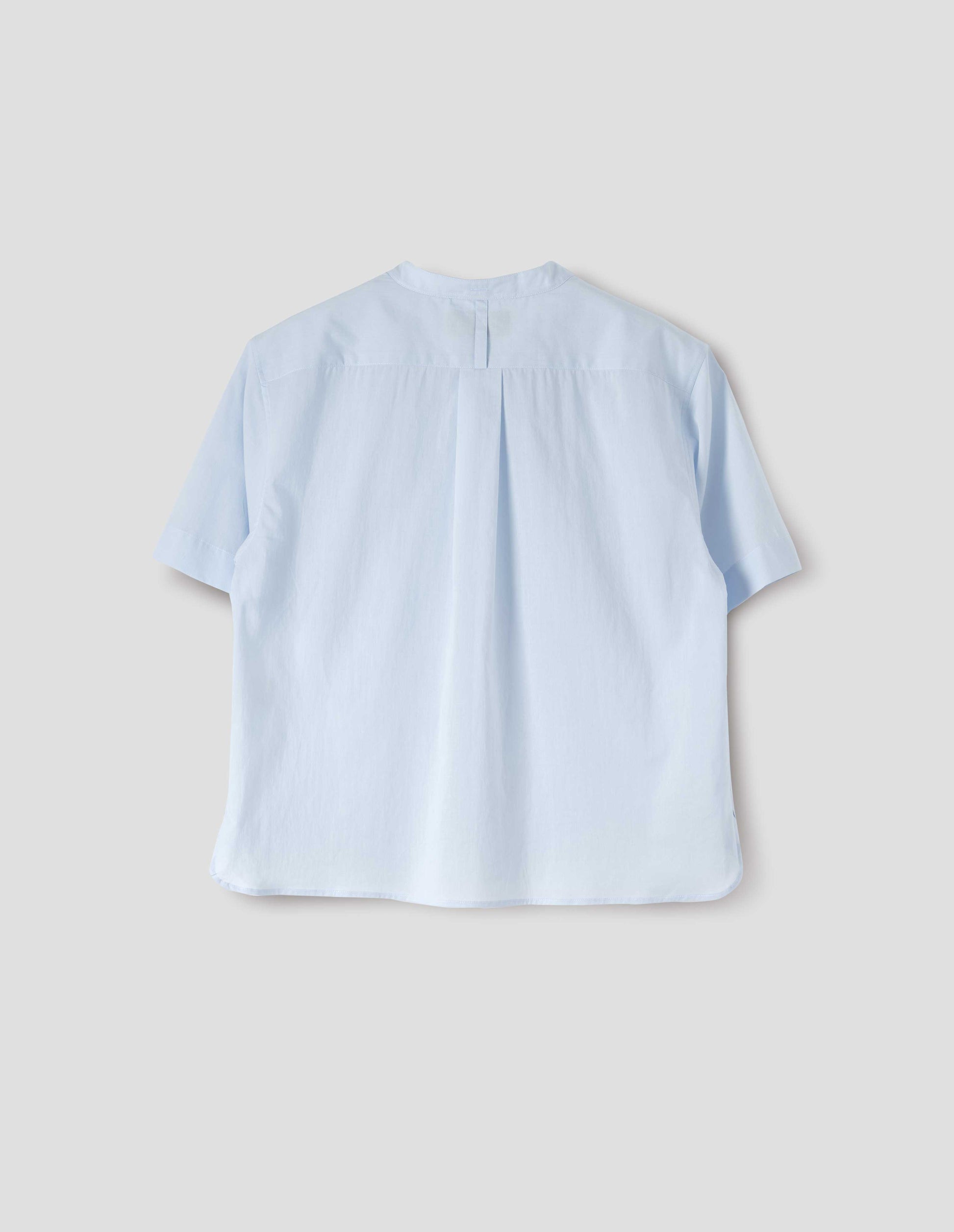 Light blue collarless shirt with short sleeves, side split stepped hem, box pleat and hanger loop detail on back yoke, and buttonhole detailing on collar band. Back flat lay view.