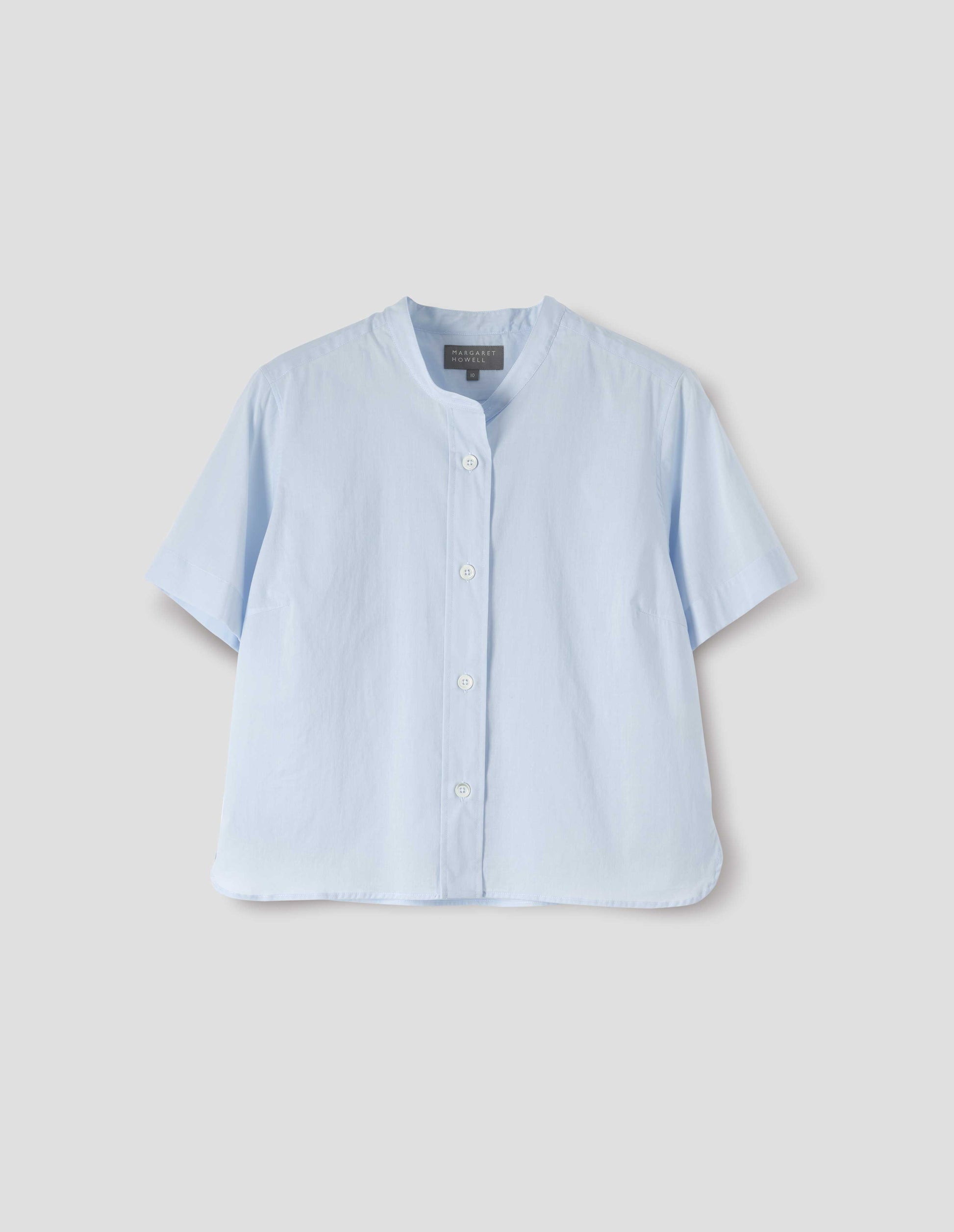 Light blue collarless shirt with short sleeves, side split stepped hem and mother of pearl buttons on raised placket. Front flat lay view.