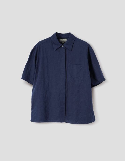 Dark blue linen short sleeve shirt, with dropped shoulder, fly front, chest pocket and side split hem. Front flat lay view.
