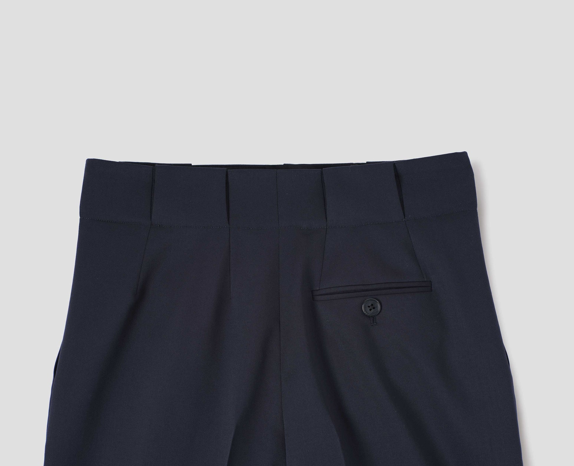 Dark navy wool trousers, grown-on waistband with large belt loops and a button-through jet back pocket and horn button. Back flat lay view.