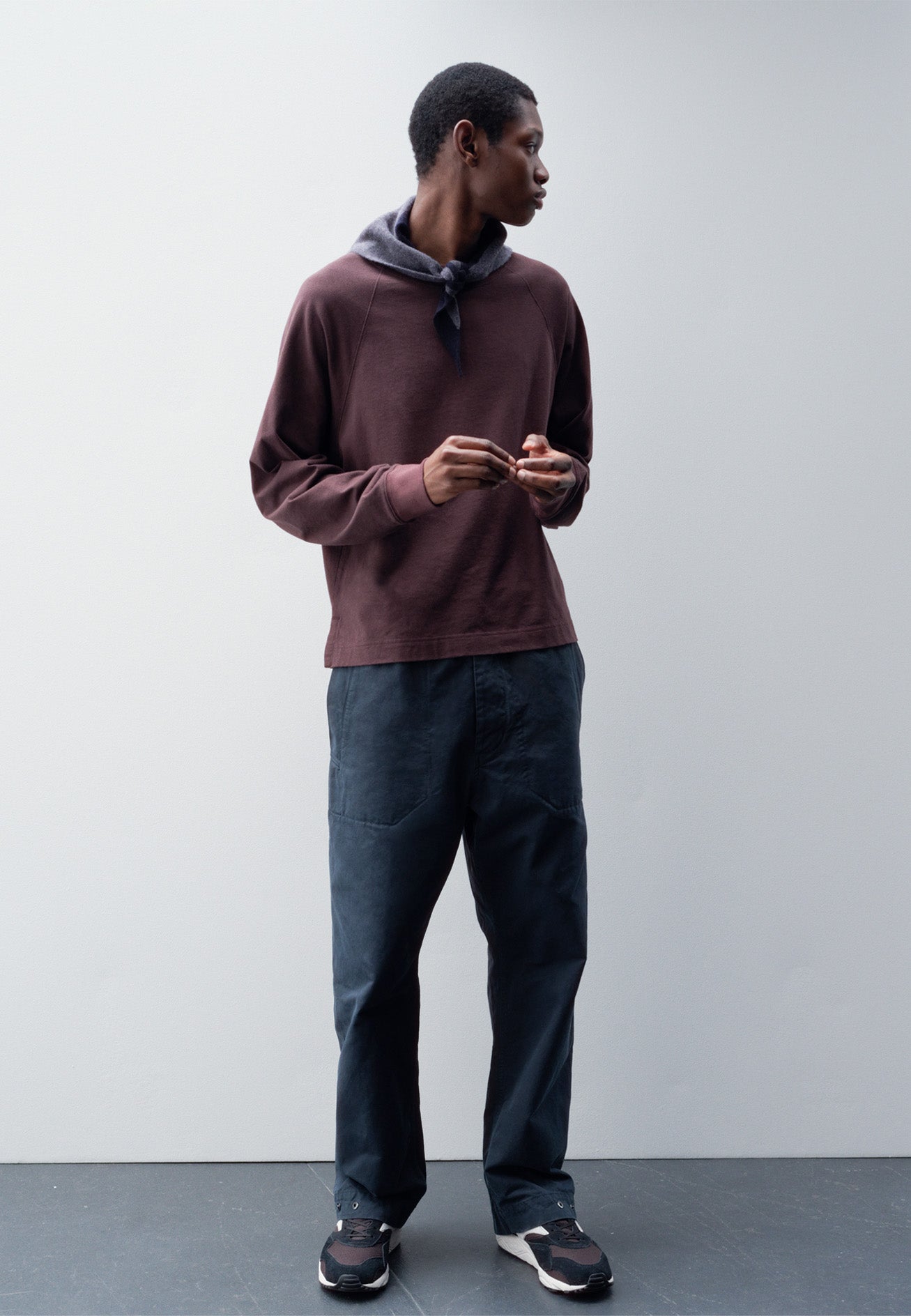Full body image, front view of model wearing relaxed blue trousers with slant welt pockets. Styled with purple long sleeve t-shirt, grey knitted scout scarf and black and brown trainers.