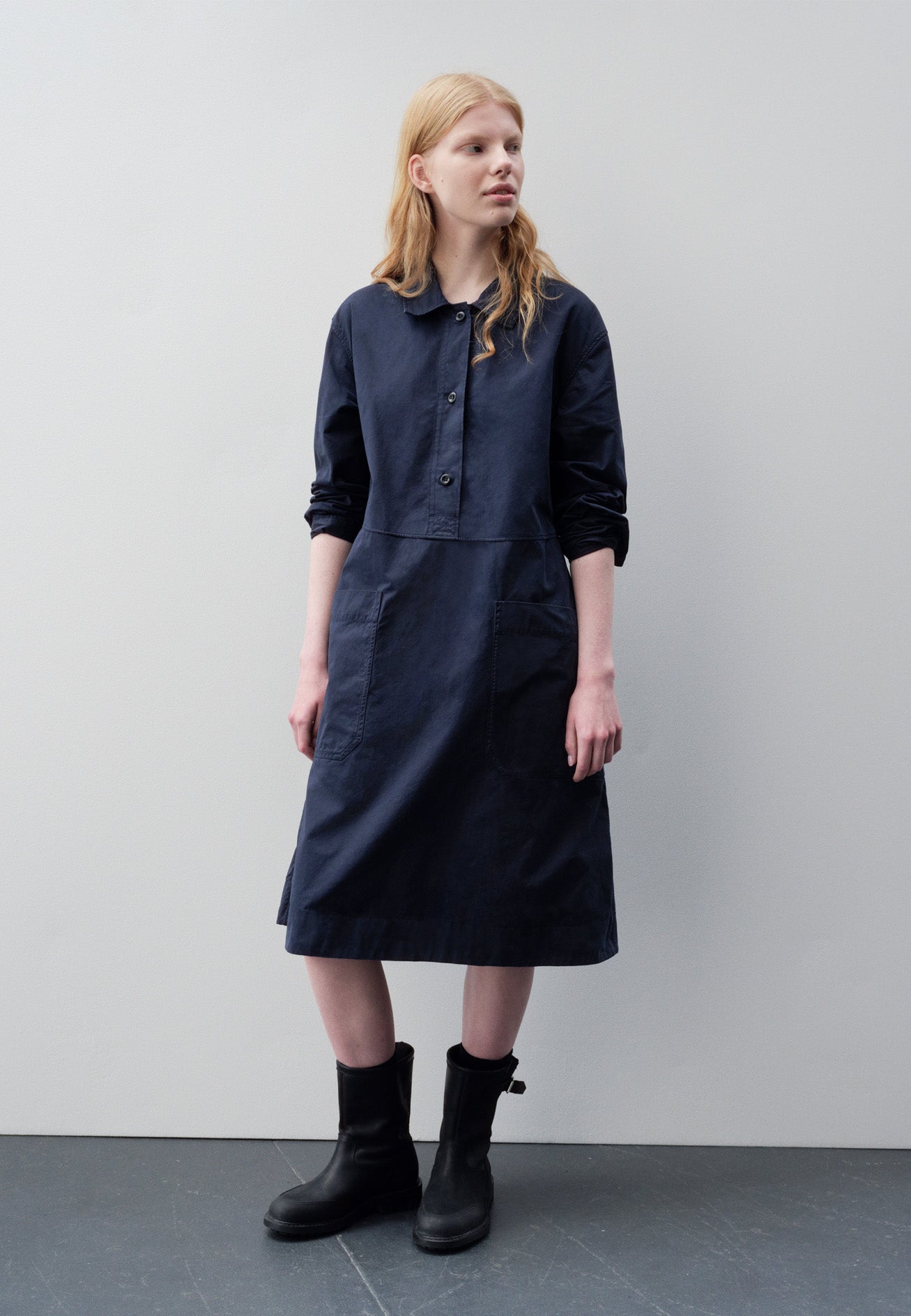 Full body image, front view of model wearing dark blue long sleeve shirt dress with box stitch detailing, large apron patch pockets, deep stitch hem and waist seam detail. Styled with black leather boots.