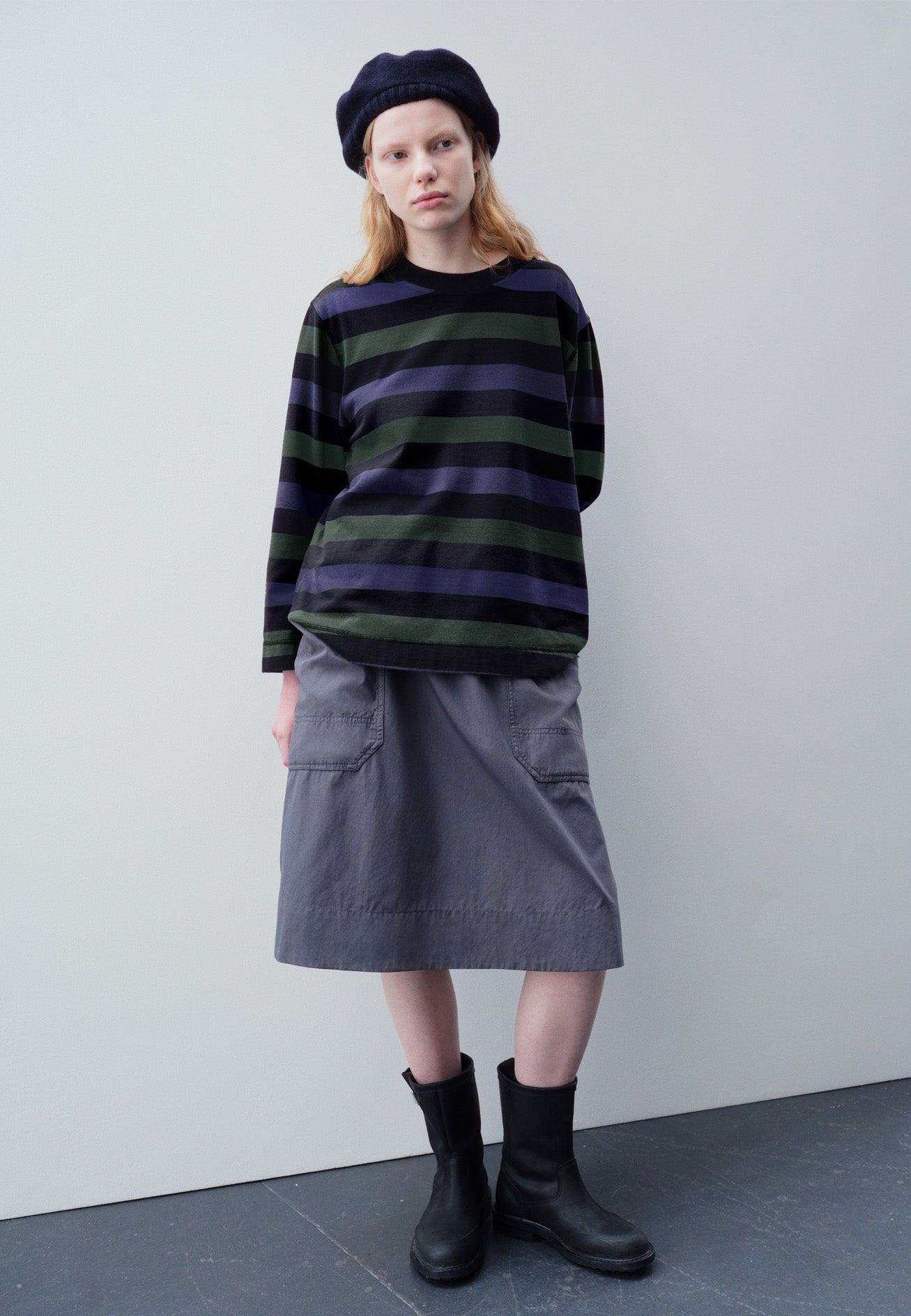 Full body image, front view of model wearing off black, blue and green striped long sleeve top, with contrast rib neck. Styled with navy knitted hat, greyish blue patch pocket skirt and black leather boots.