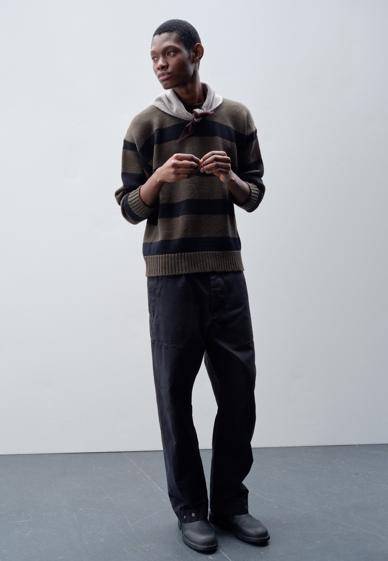 Full body image, front view of model wearing green and black striped wool jumper, with ribbed neck, cuffs and hem. Styled with grey knitted scout scarf, black trousers and black leather boots.