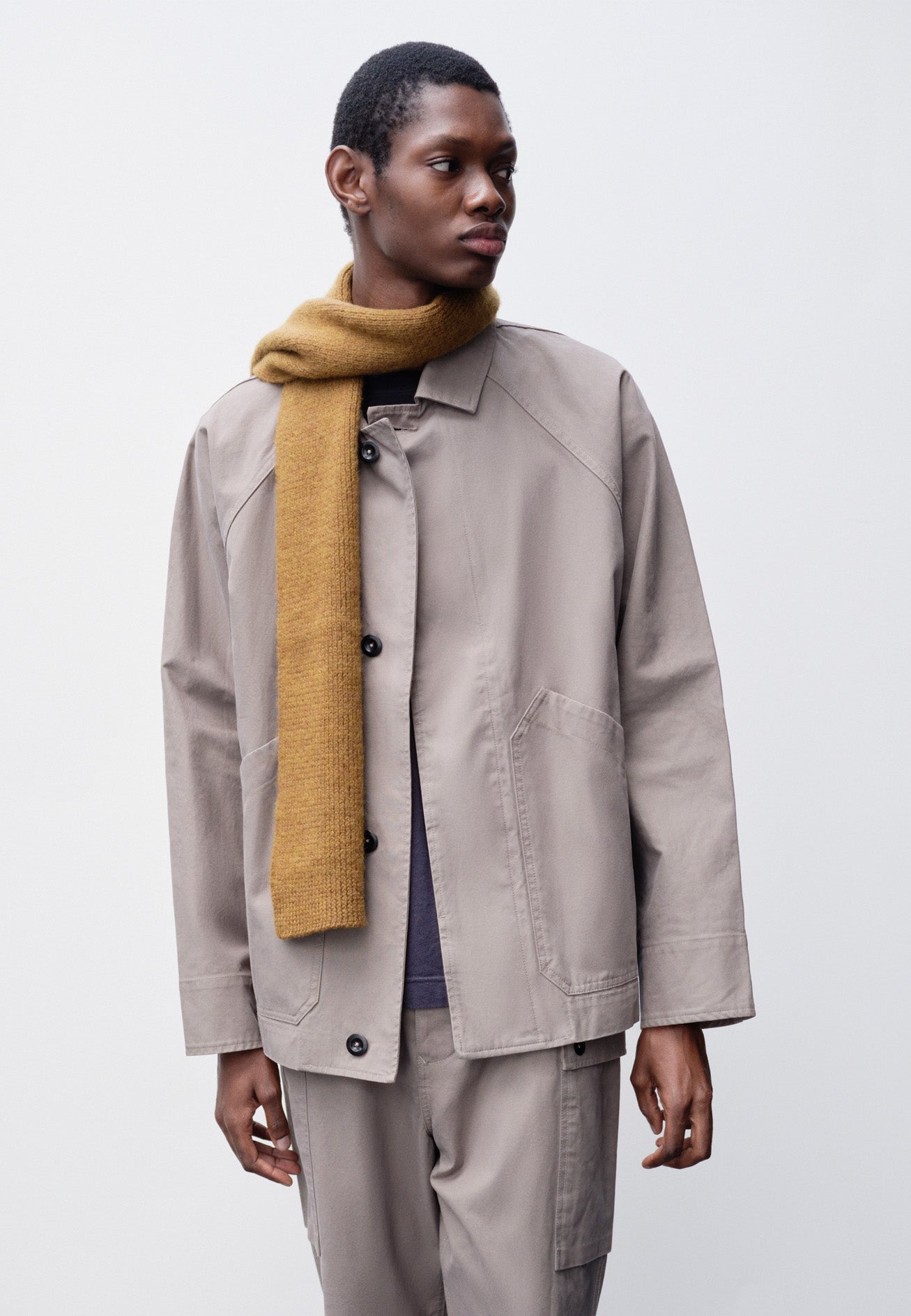 Cropped body image, front view of model wearing beige raglan sleeve jacket with corozo button fastening fly front and slant patch pockets. Styled with matching trousers, navy blue t-shirt and yellow knitted scarf.