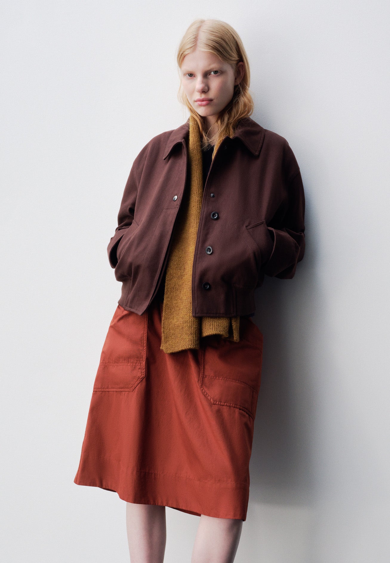 Full body image, front view of model wearing dark orange relaxed midi A-line skirt with elasticated waist and large, slant patch pockets. Styled with brownish red collared jacket and yellow knitted scarf.