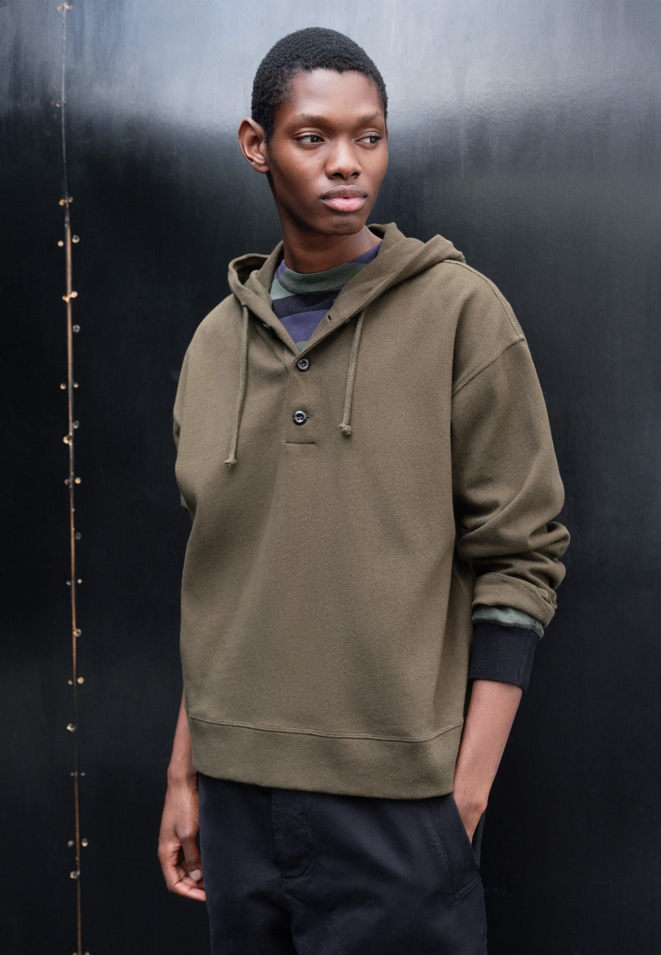 Cropped body image, front view of model wearing green cotton hoodie, with drawcord hood, corozo buttons, straight cuffs and stitch seam detailing. Styled over green, black and blue striped long sleeve t-shirt and with navy blue trousers.