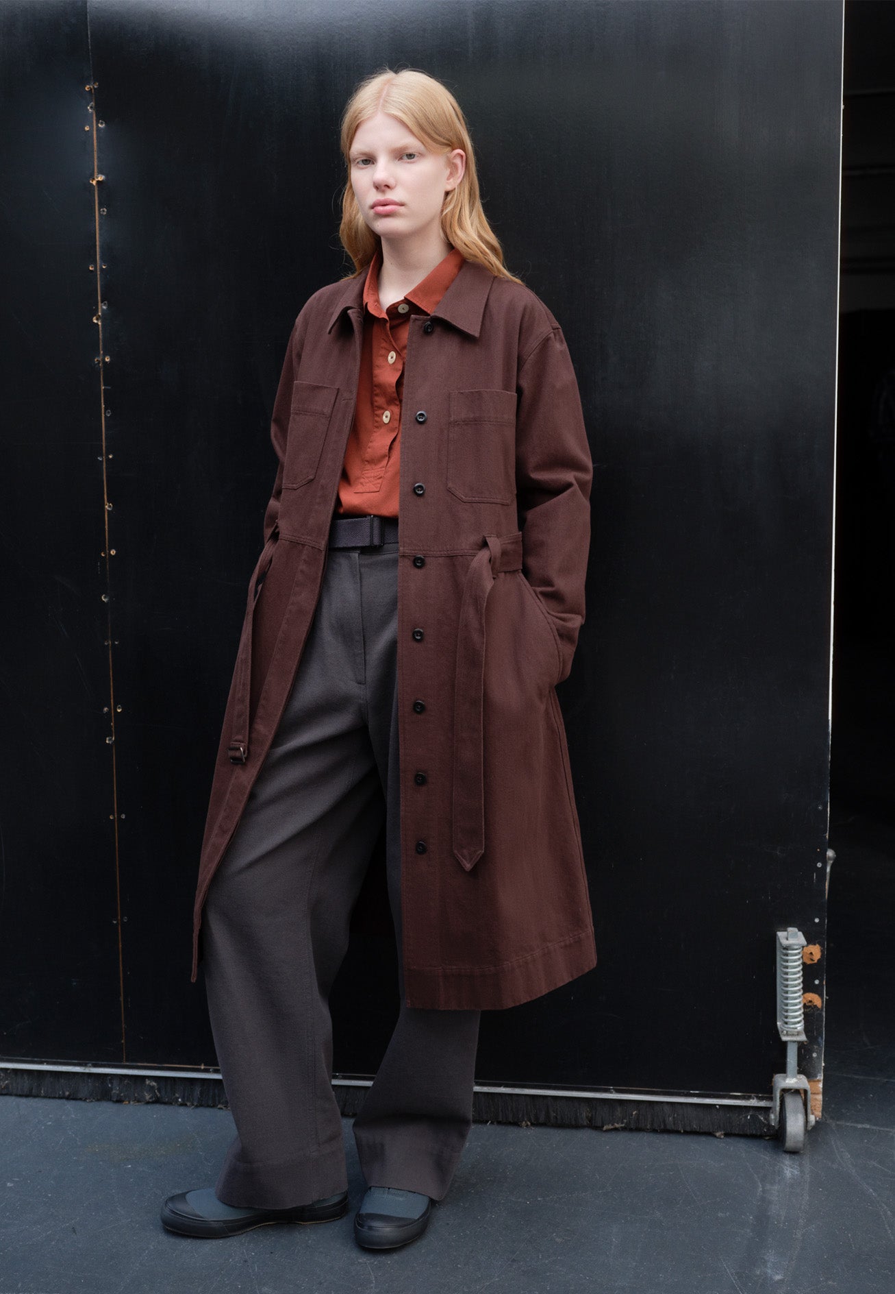 Full body image, front view of model wearing brown red long sleeve shirt dress, with fly front corozo button fastening, patch chest and slant side pockets, button fastening cuffs and removeable belt with metal slide adjuster. Styled open like a coat with red shirt and grey belted trousers underneath, with blue canvas trainers.