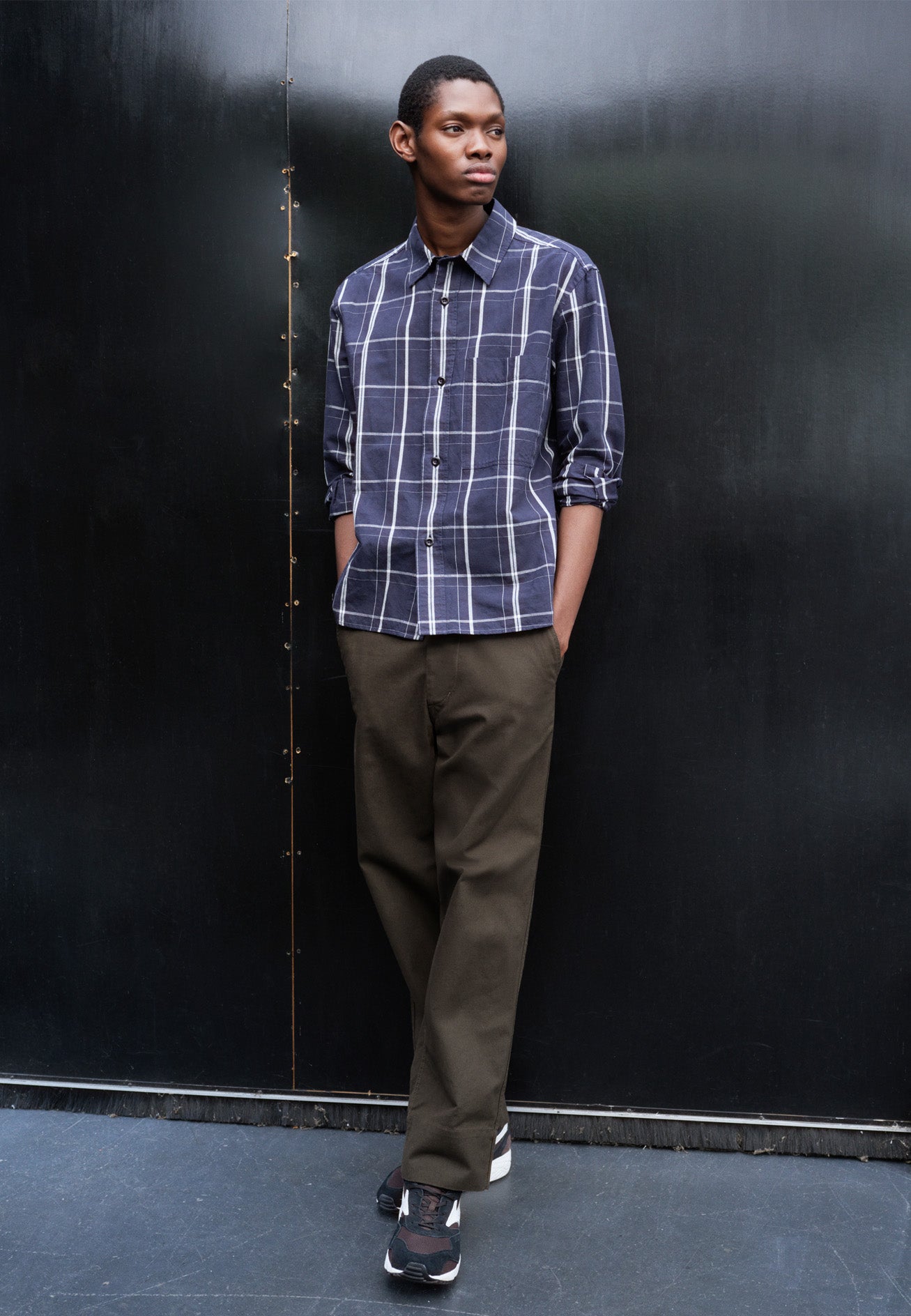 Full body image, front view of model wearing blue and white checked long sleeve shirt. With raised placket, corozo brace buttons and button through chest patch pocket. Styled with green trousers and black and brown trainers.