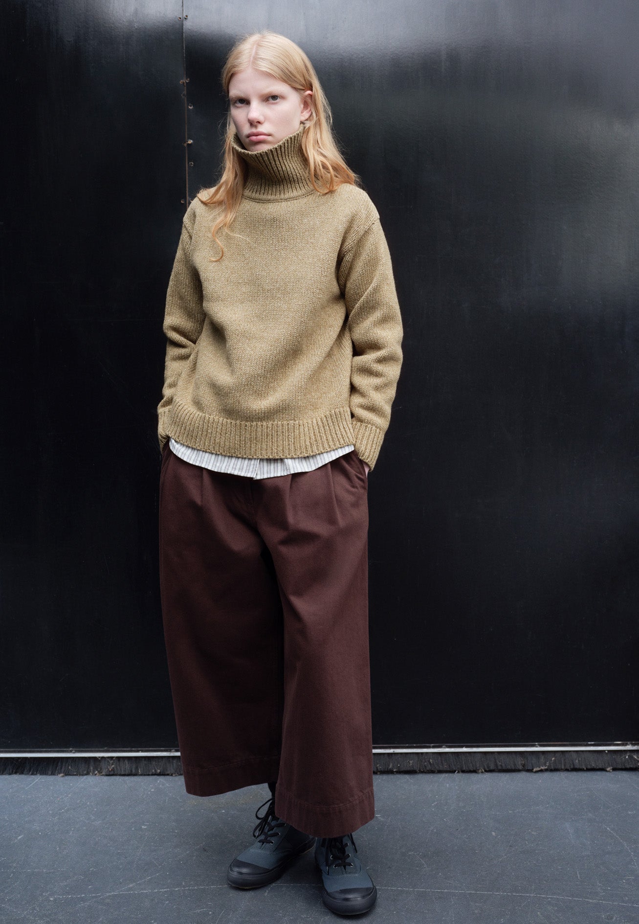 Full body image, front view of model wearing red brown double pleated culottes, with button and zip fastening, belt loops and side seam pockets. Styled with green high neck jumper, white and grey striped shirt and blue high top canvas trainers.