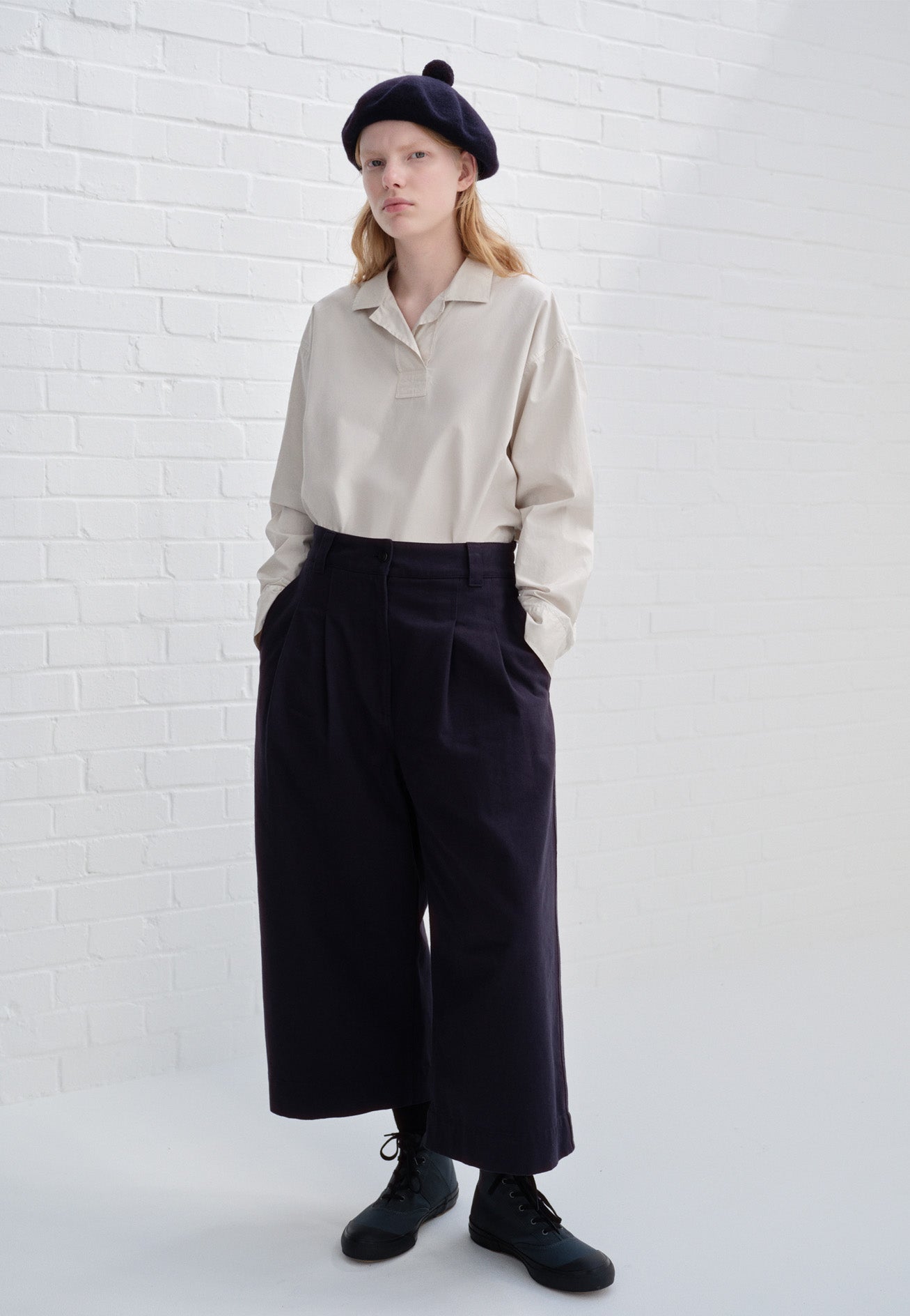 Full body image, front view of model wearing dark blue double pleated culottes, with button and zip fastening, belt loops and side seam pockets. Styled with off white shirt, navy blue bobble hat and blue high top canvas trainers.