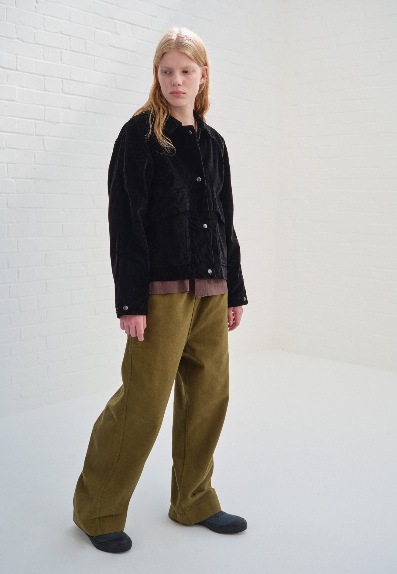 Full body image, front view of model wearing green relaxed wide leg joggers. Styled with black corduroy jacket, purple long sleeve top and blue canvas trainers.