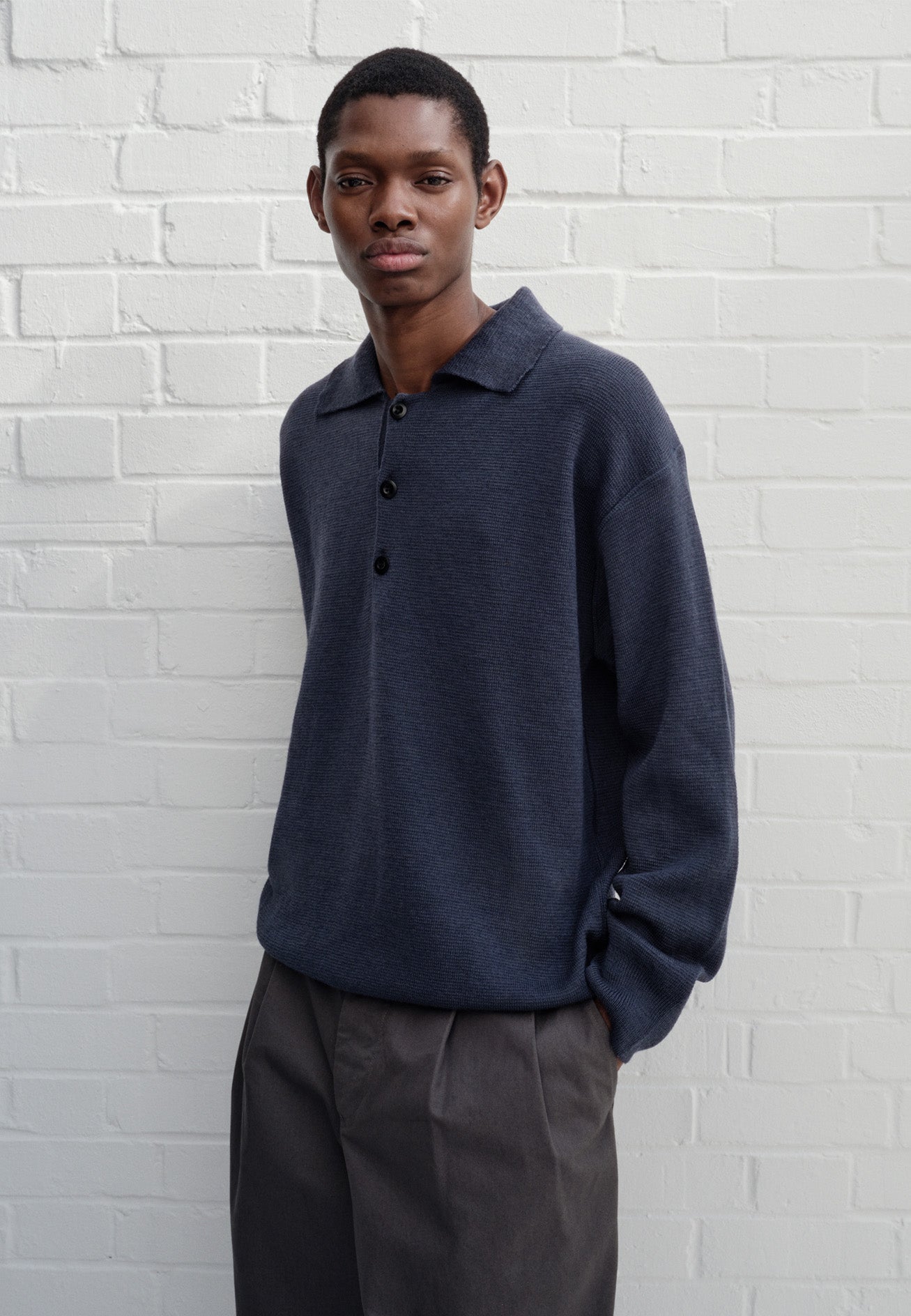 Full body image, front view of model wearing blue knitted polo with contrast rib on neck, cuffs and hem and corozo brace buttons. Styled with grey pleated trousers.
