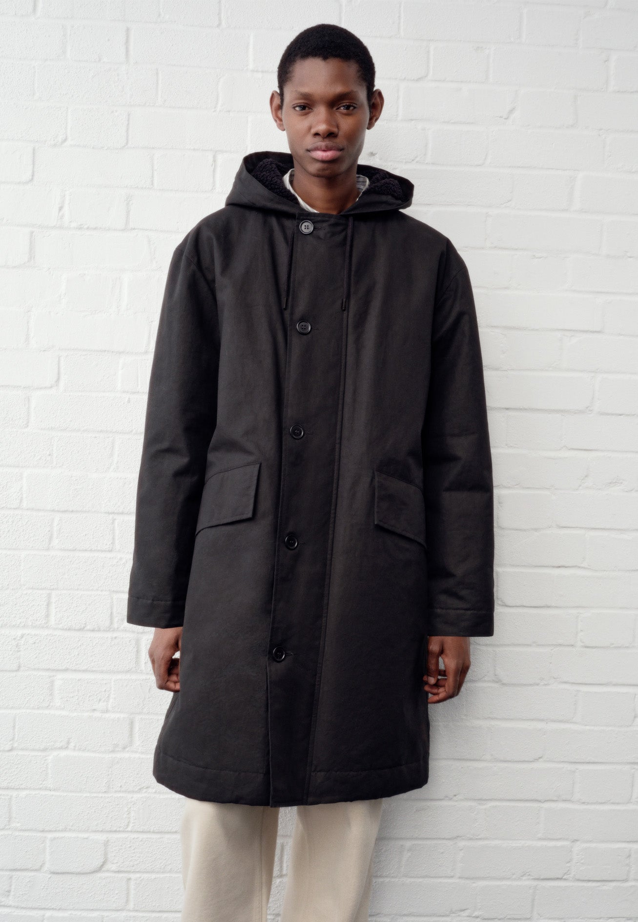 Full body image, front view of model wearing black hooded parka with fleece lined drawcord hood, two way zip fastening with button fastening storm flap and slanted flap pockets. Styled over beige trousers.