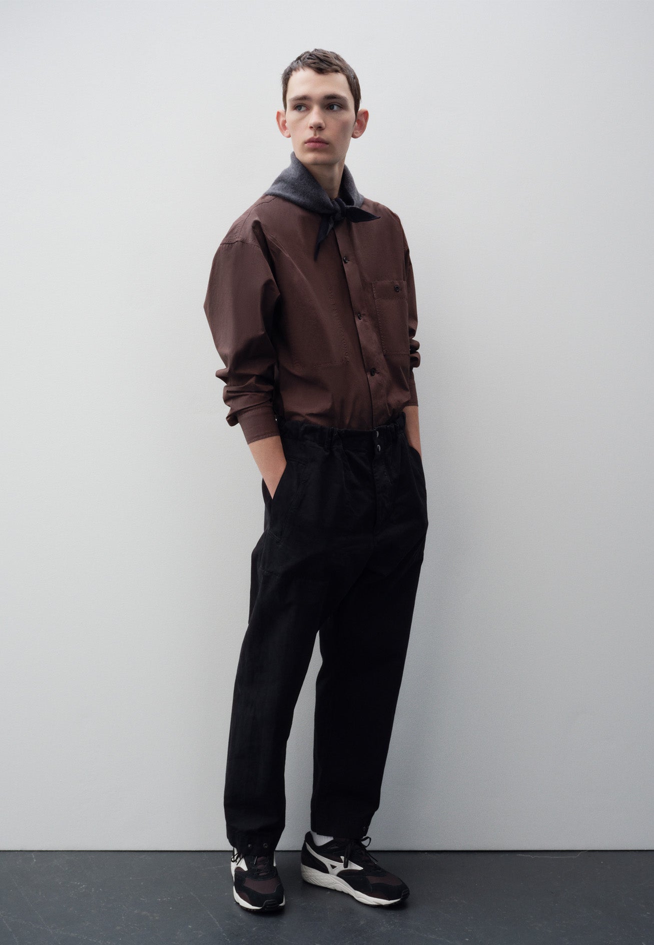 Full body image, front view of model wearing relaxed black trousers with slant welt pockets, corozo brace button fly and press stud hem adjusters. Styled with reddish brown collarless shirt, grey knitted scout scarf and black and brown trainers.