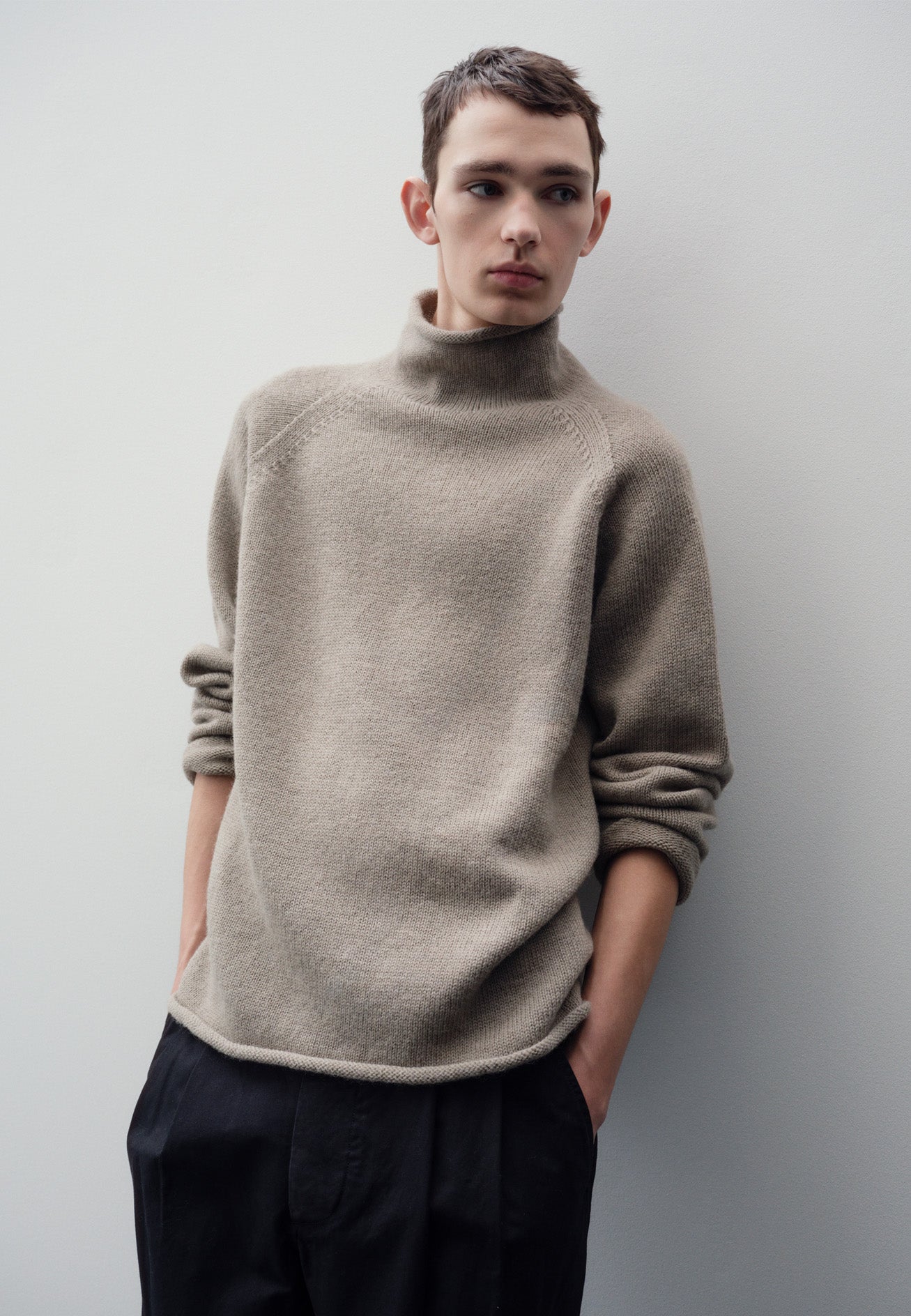 Cropped body image, front view of model wearing beige roll neck jumper with seamless construction, high neck, raglan sleeve and straight cuffs and hem. Styled with black trousers.