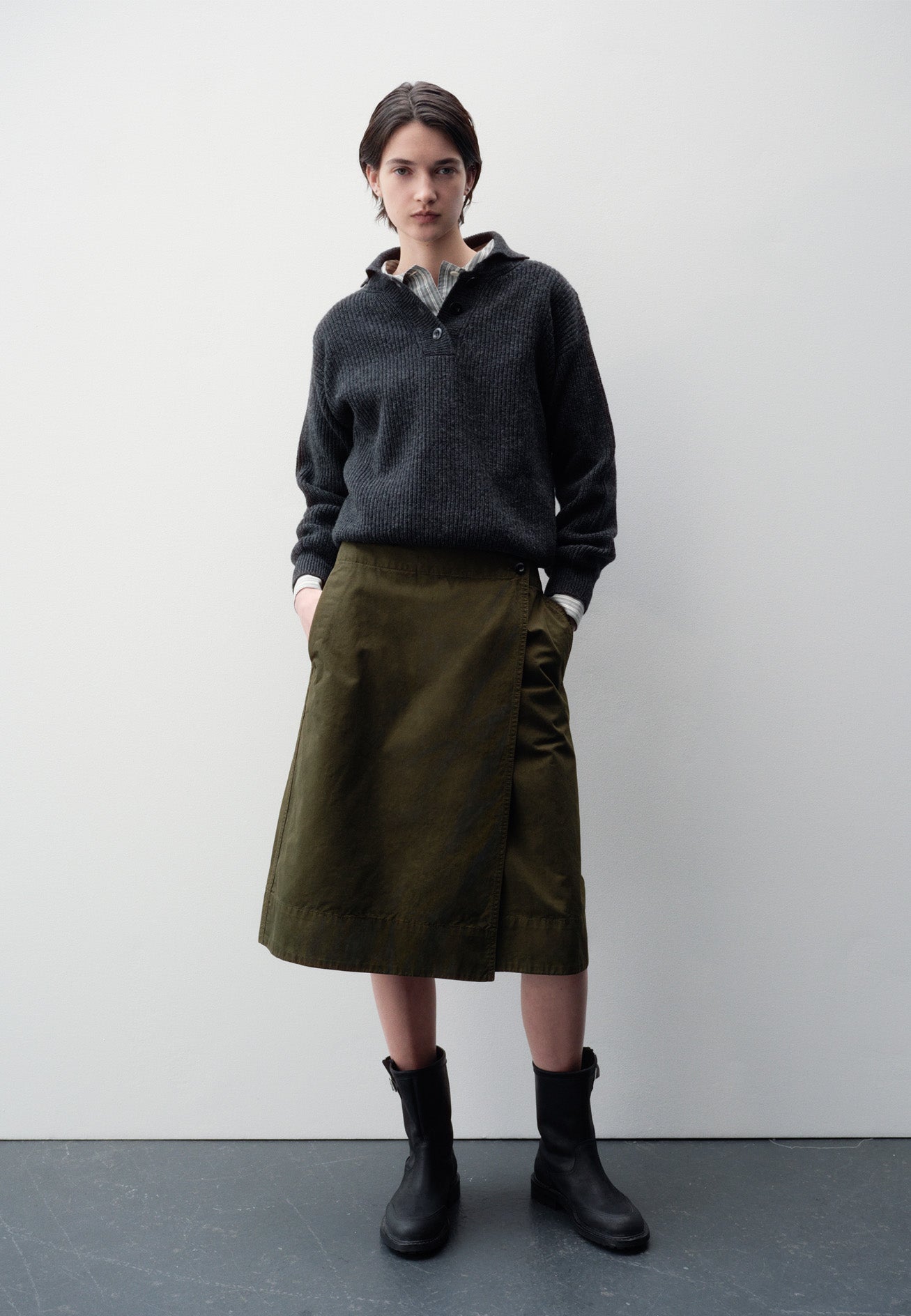 Full body image, back view of model wearing green skirt with deep wrap and slant side pockets. Styled with grey and white striped shirt, grey knitted polo and black leather boots.