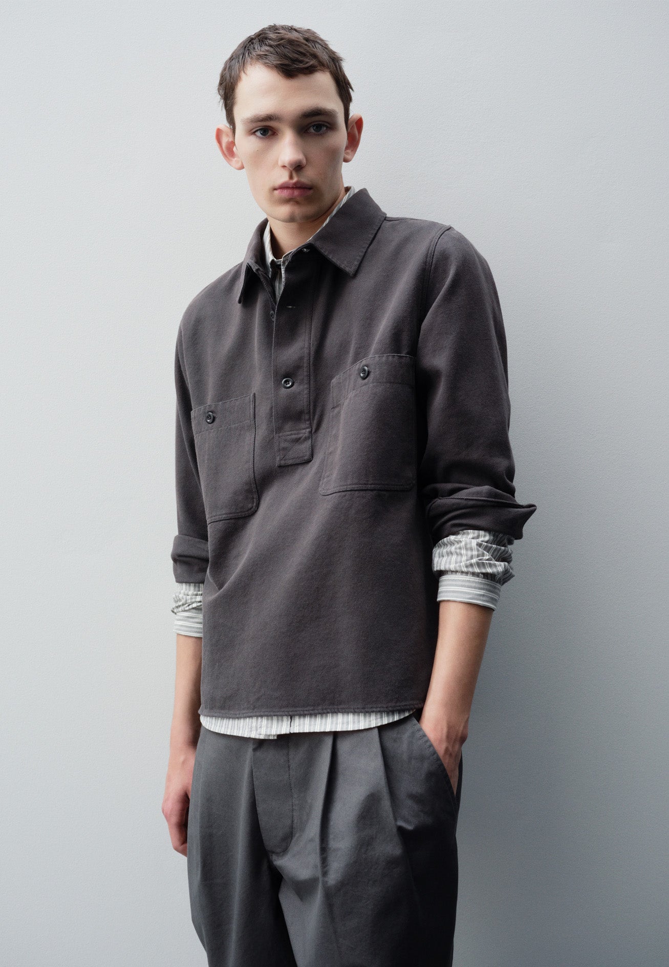 Cropped body image, front view of model wearing grey overhead shirt with raised placket, box stitch detailing, button through chest patch pockets and elongated back hem. Styled over green, grey and white striped shirt and grey pleated trousers.