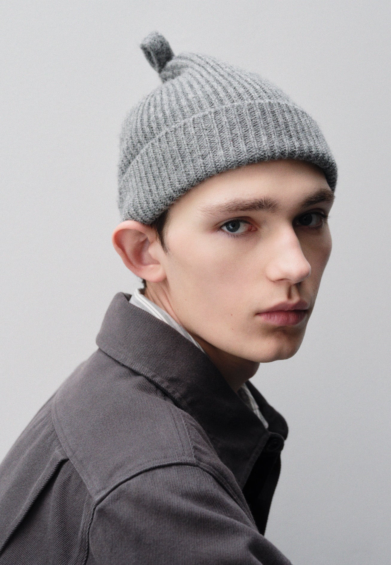 Cropped image, side view of male model wearing grey wool ribbed hat, with knitted loop detailing on top. Styled with grey shirt. 