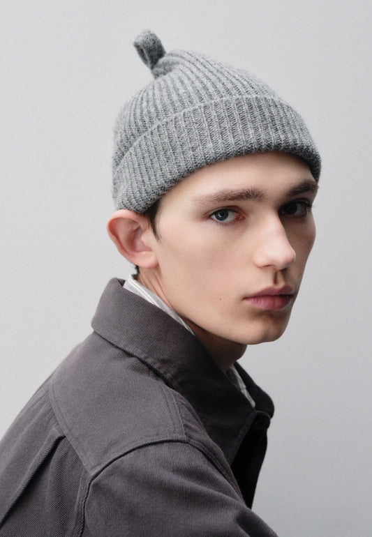 Cropped image, side view of male model wearing grey wool ribbed hat, with knitted loop detailing on top. Styled with grey shirt. 