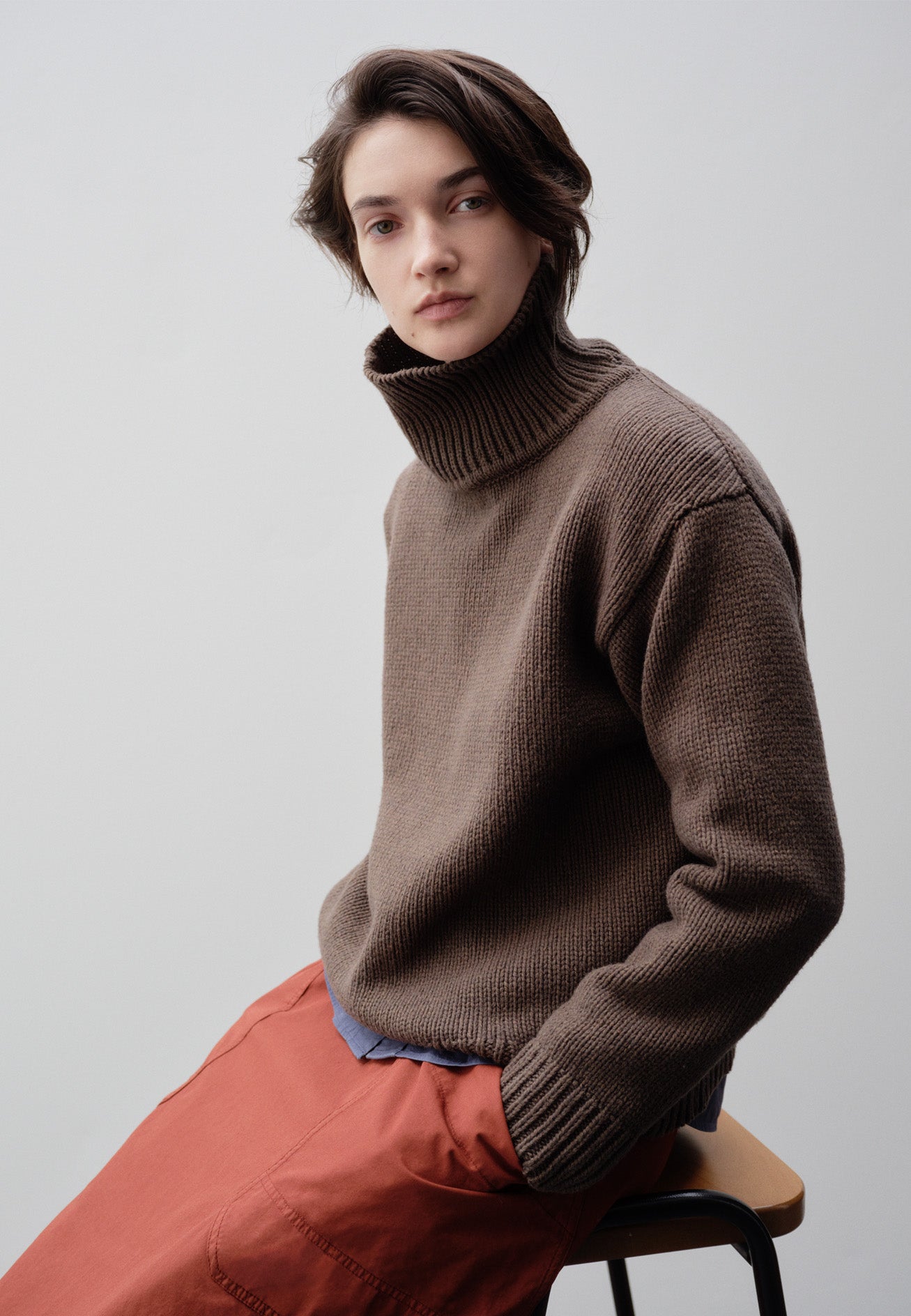Cropped image, front view of model sat down wearing relaxed brown high neck jumper with deep rib trim on neck, cuffs and hem. Styled with red patch pocket skirt.
