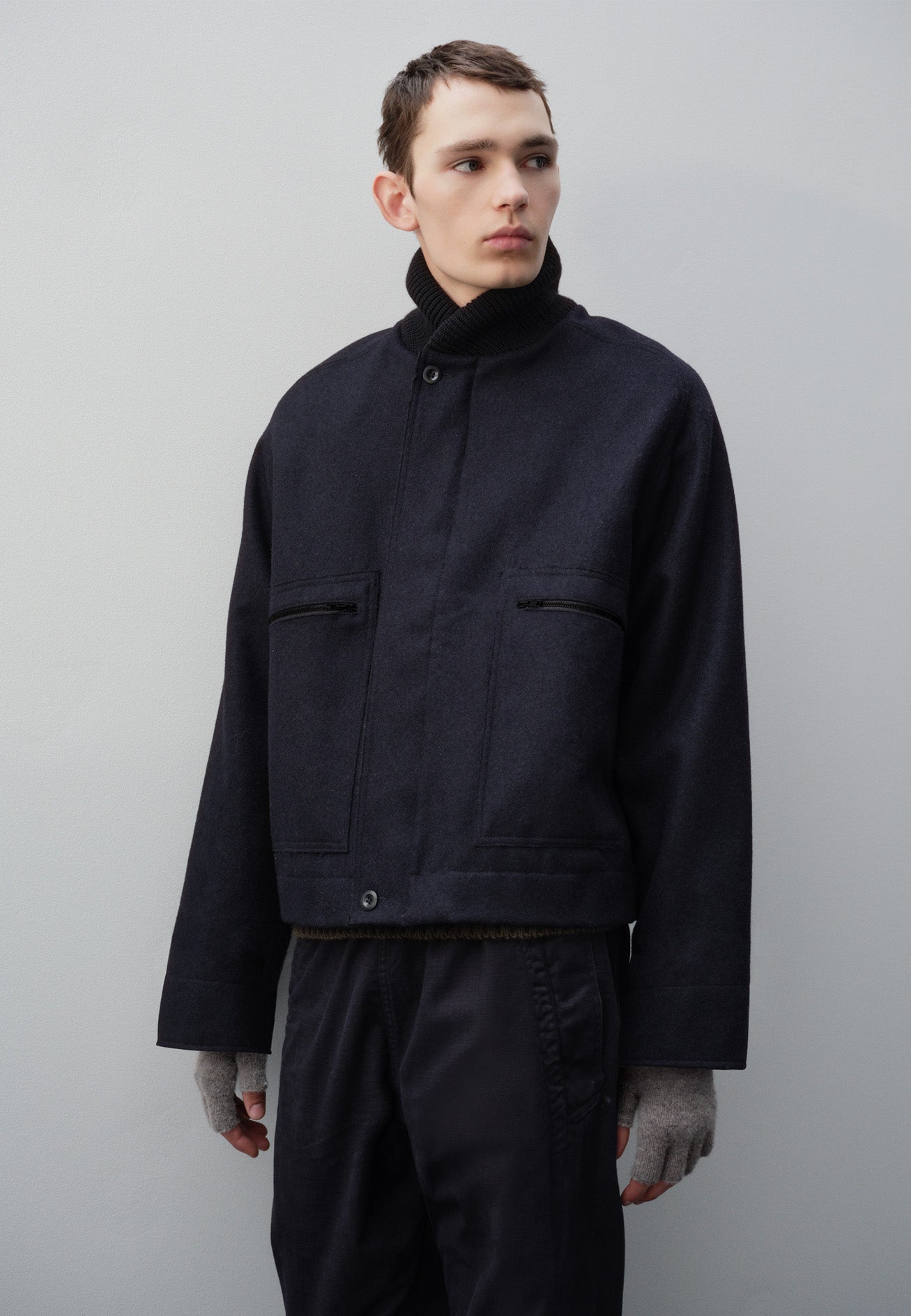Cropped body image, front view of model wearing navy blue rib neck jacket, with contrast wool rib collar, saddle sleeves, button through cuffs, patch zip pockets and raw edge detailing. Styled with indigo blue trousers and grey fingerless gloves.