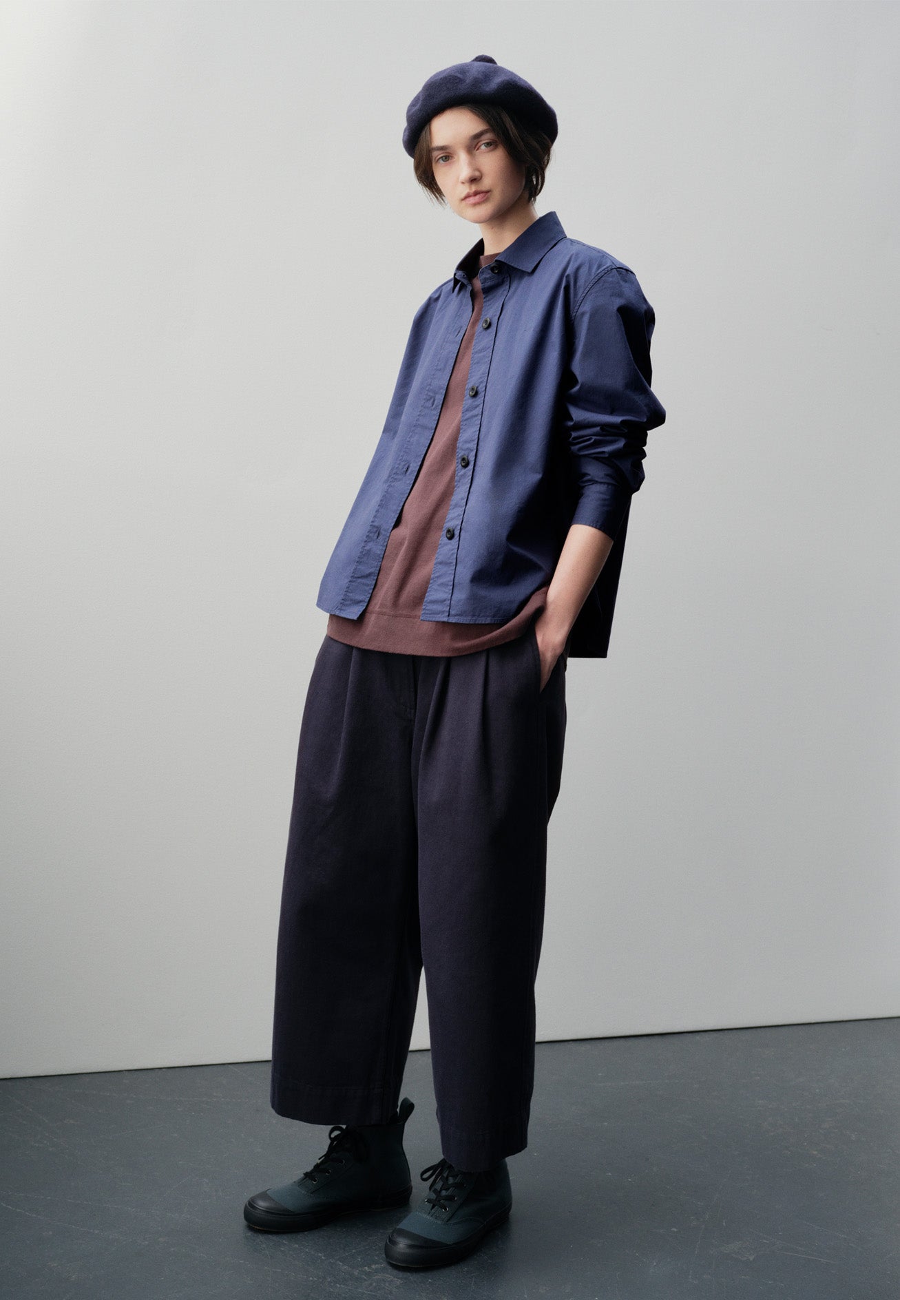 Full body image, front view of model wearing indigo blue shirt with raised placket and corozo button closure. Styled over purple long sleeve t-shirt, with navy bobble hat, navy blue culottes and blue high top canvas trainers. 