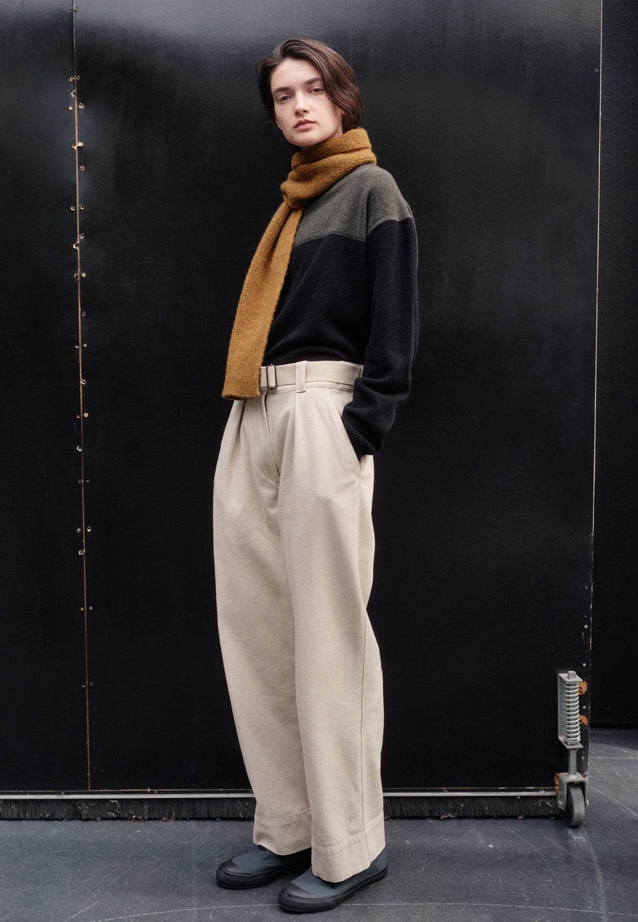 Full body image, side view of female model wearing yellow knitted wool scarf. Styled with black and green jumper, off white trousers and blue canvas trainers.