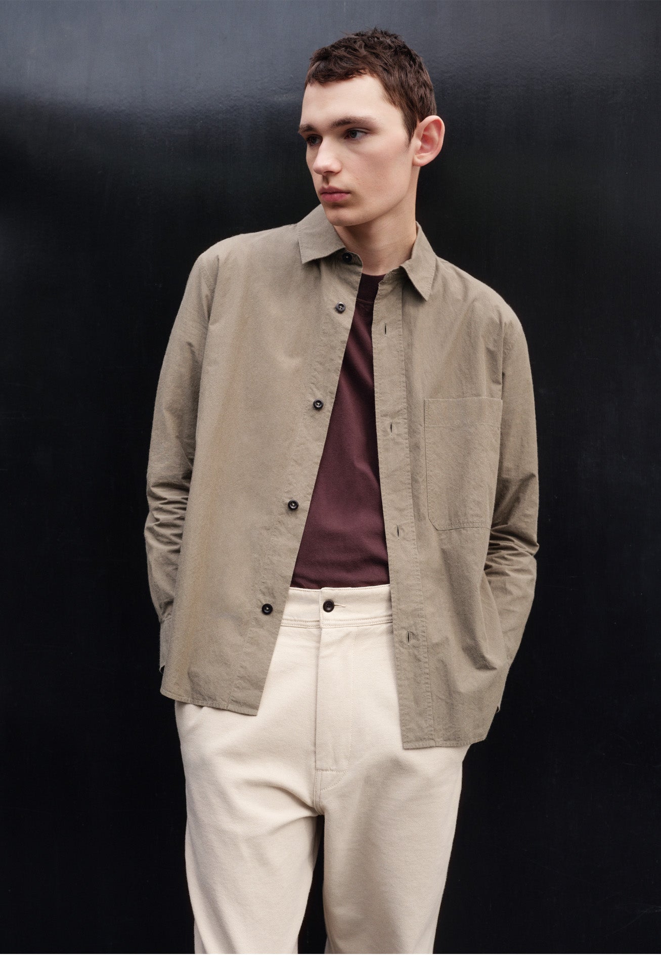 Cropped body image, front view of model wearing green brushed cotton shirt with raised placket, large chest pocket, corozo button closure and side gusset stitch detailing. Styled over purple t-shirt with off white trousers.