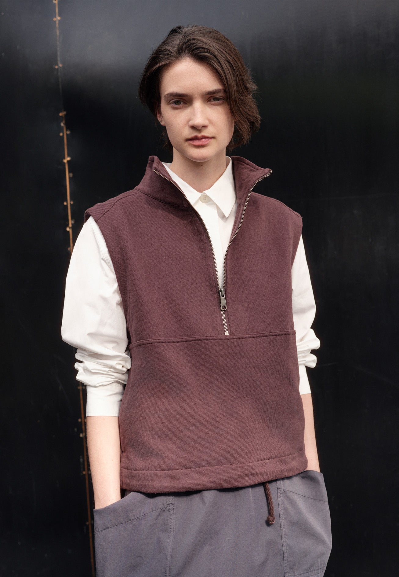 Cropped image, front view of model wearing purple brown half zip high neck sleeveless track top and drawcord hem. Styled with white cotton shirt and greyish blue patch pocket skirt.