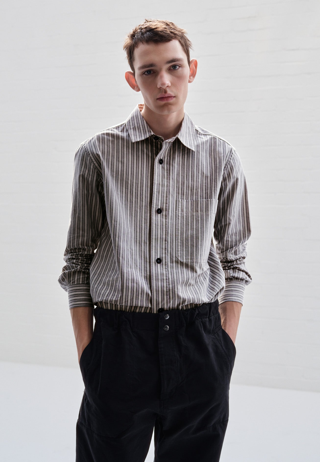 Cropped body image, front view of model wearing green, grey and white striped cotton long sleeve shirt, with raised placket, large chest pocket, corozo button closure and side gusset stitch detailing. Styled with black trousers.