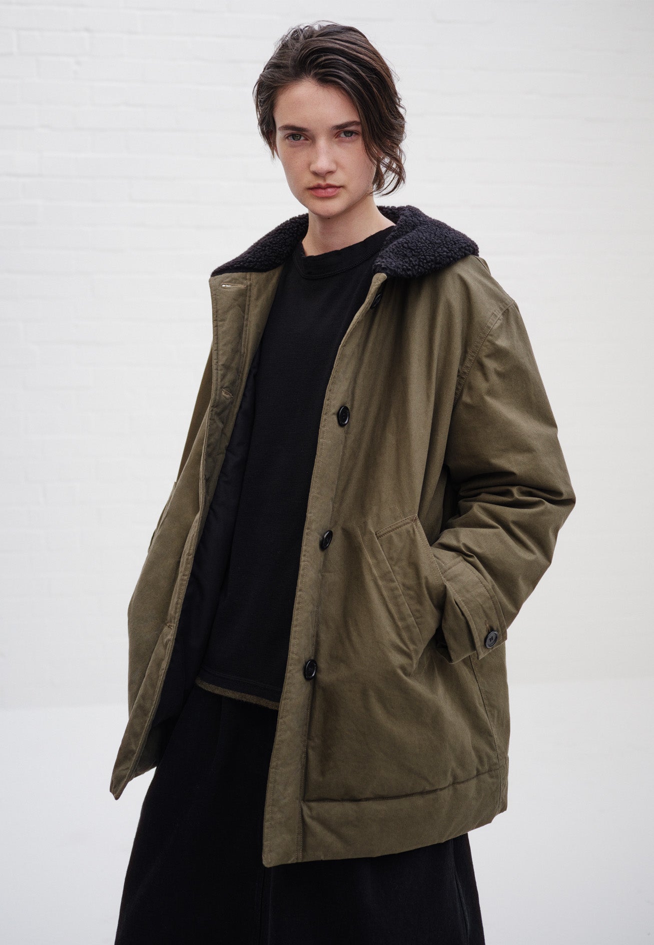 Cropped image, front view of model wearing relaxed khaki green coat with black fleece collar, slanted welt pockets, corozo brace buttons and button tab cuff adjusters. Styled over black jumper and black skirt.