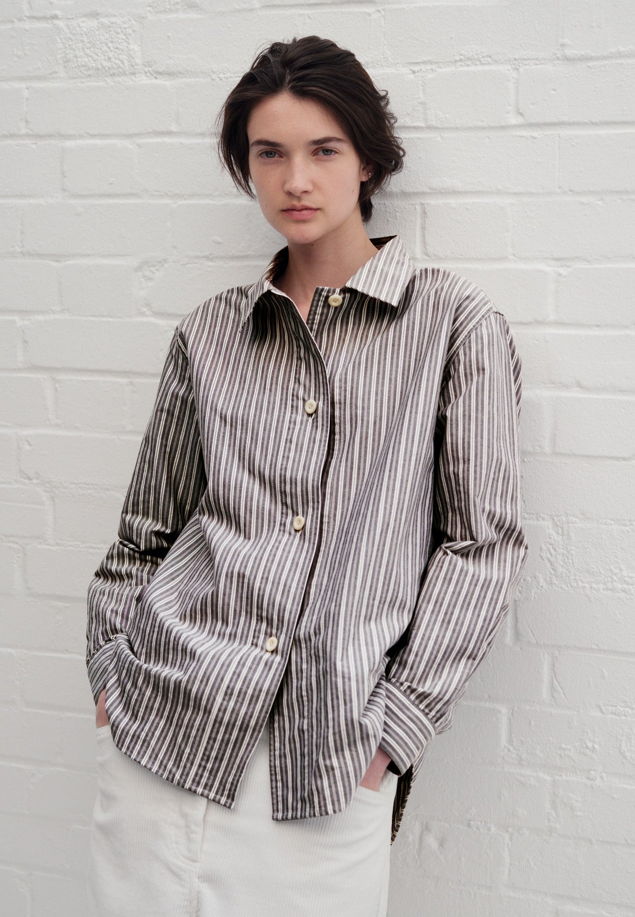 Cropped body image, front view of model wearing green, grey and off white striped long sleeve shirt. With wide stitch through placket and corozo brace buttons and cuffs. Styled with white trousers.