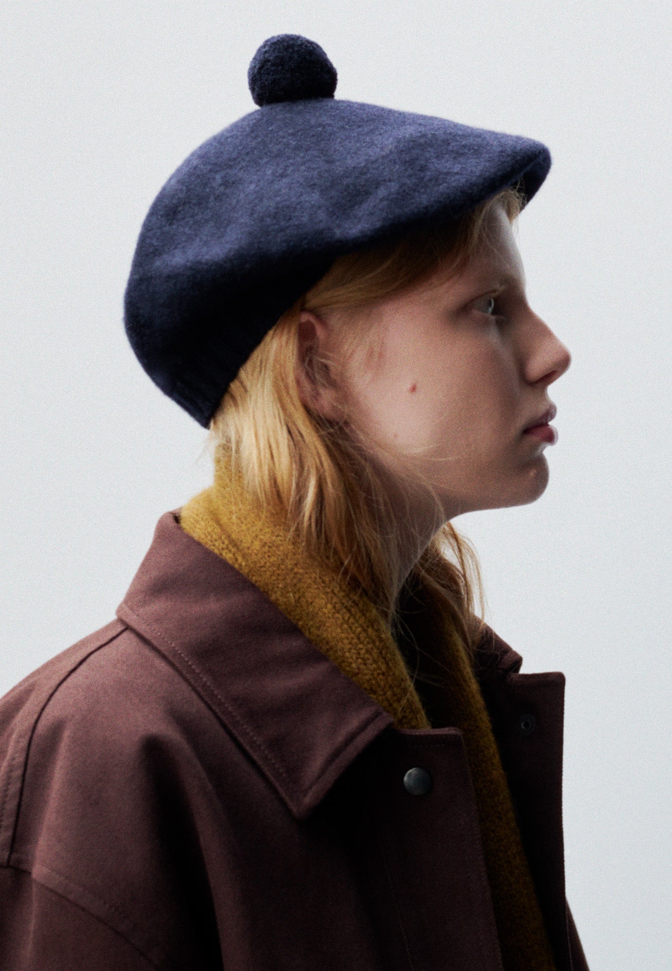 Cropped body image, side view of female model wearing dark navy wool tam o shanter hat with matching pom pom bobble in centre. Styled with yellow wool scarf and burgundy jacket.
