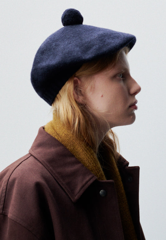 Cropped body image, side view of female model wearing dark navy wool tam o shanter hat with matching pom pom bobble in centre. Styled with yellow wool scarf and burgundy jacket.