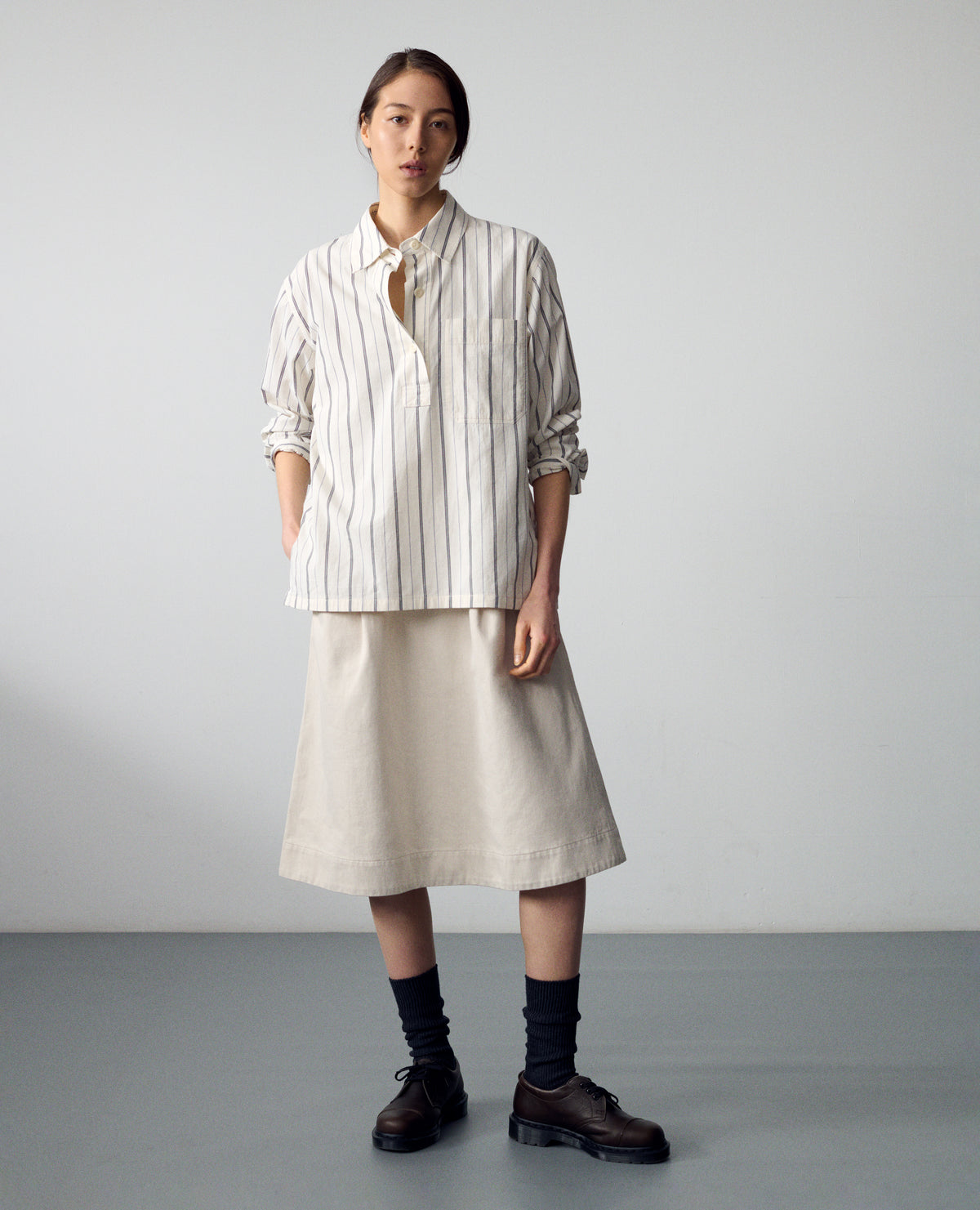 Full body image, front view of model wearing an off white and dark blue stripe cotton shirt with light buttons, a half placket and a chest patch pocket. Styled over an off white cotton drill dress, black socks and brown leather shoes.