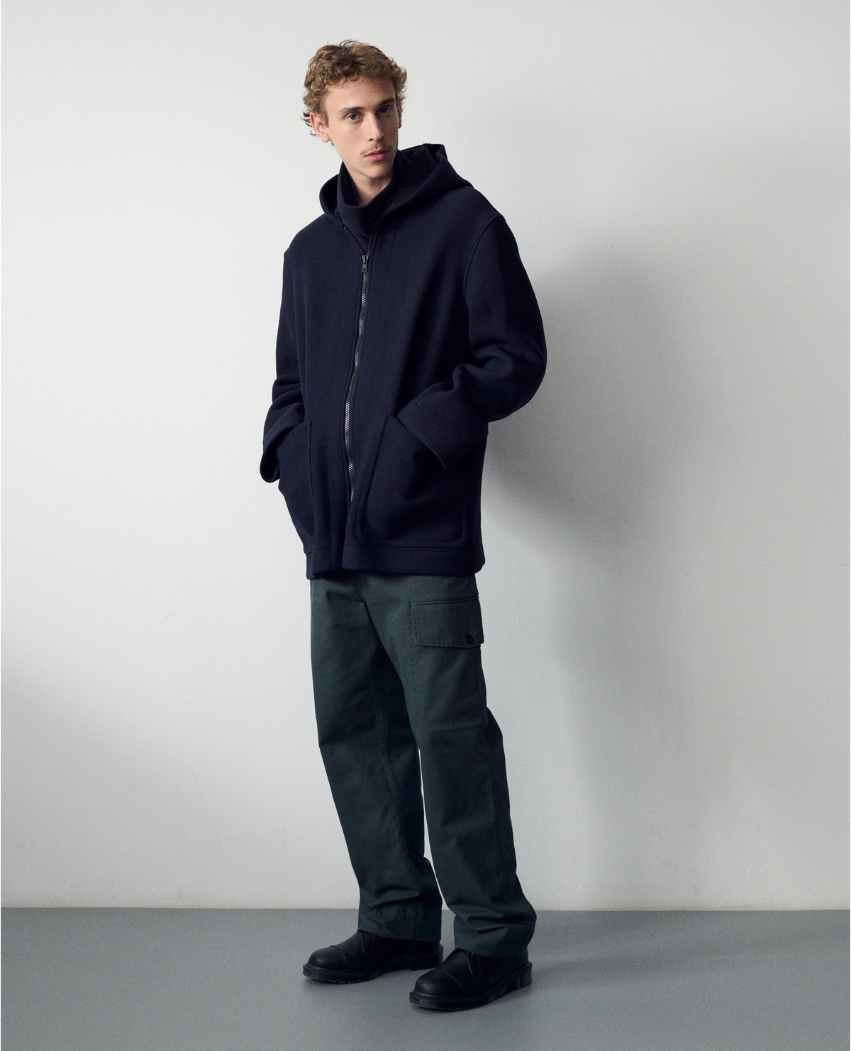 Full body image, front view of model wearing a dark navy zip through wool coat, with a hood, two laid-on patch pockets, straight cuffs and hems. Worn with grey cargo trousers and with black leather shoes.