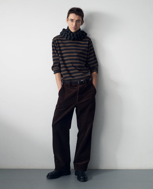 Full body image, front view of model wearing a black and brown stripe cotton jersey top. Styled with a black and brownish green knitted wool striped scarf, brown corduroy flat front trousers, black leather belt and shoes.