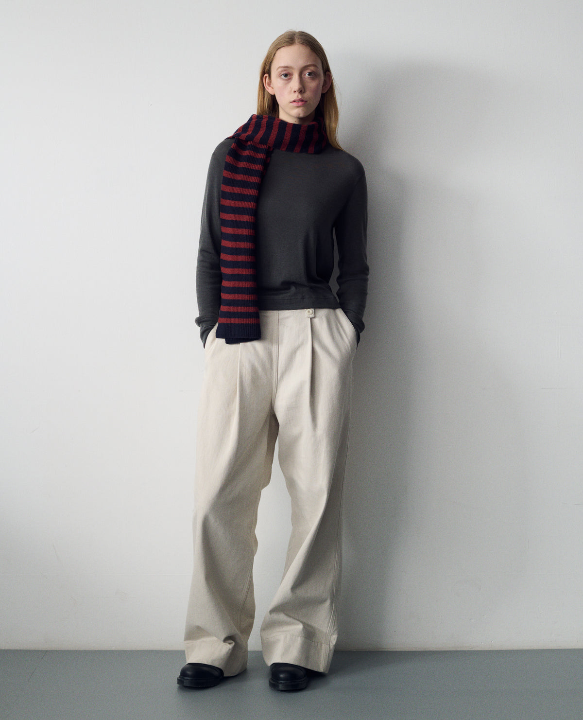 Full body image, front view of model wearing a dark grey long sleeve t shirt with set-in sleeves, dark blue and red stripe wool scarf and off white trousers with with button-fastening belt loops, a side opening with integrated side seam pockets, and two front pleats. Worn with black leather shoes.