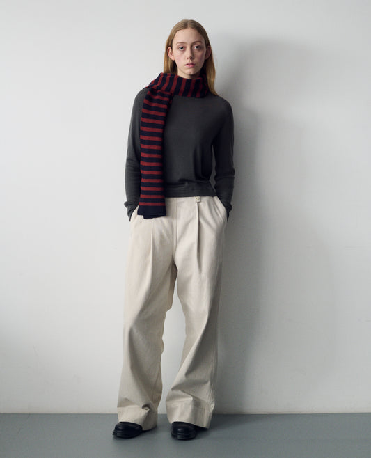 Full body image, front view of model wearing a grey wool cotton slipover with a high rib neck, rib armholes and hem. Styled over a mid length dark grey pull on cotton dress with a box pleat skirt and with black socks and leather shoes.