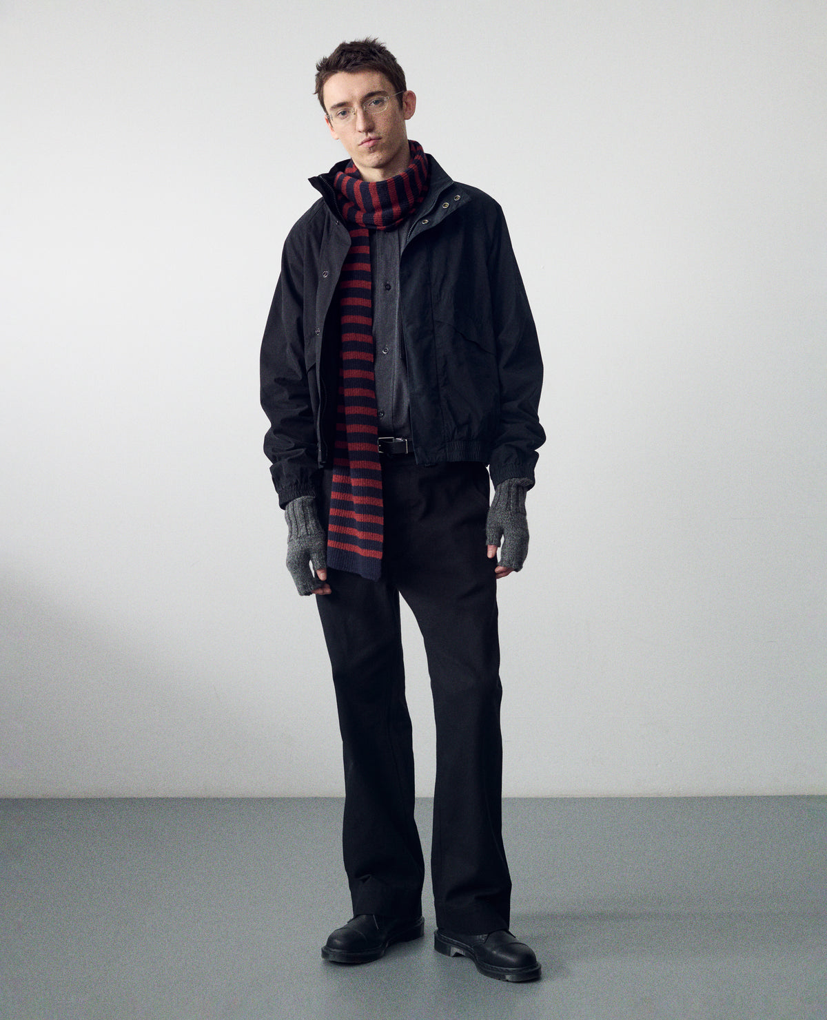 Full body image, front view of model wearing black cotton trousers with belt loops, button fastening and slanted side entry pockets. Styled with black jacket over dark grey shirt, knitted red and black striped knitted scarf, grey fingerless mittens, black leather belt and black leather shoes.