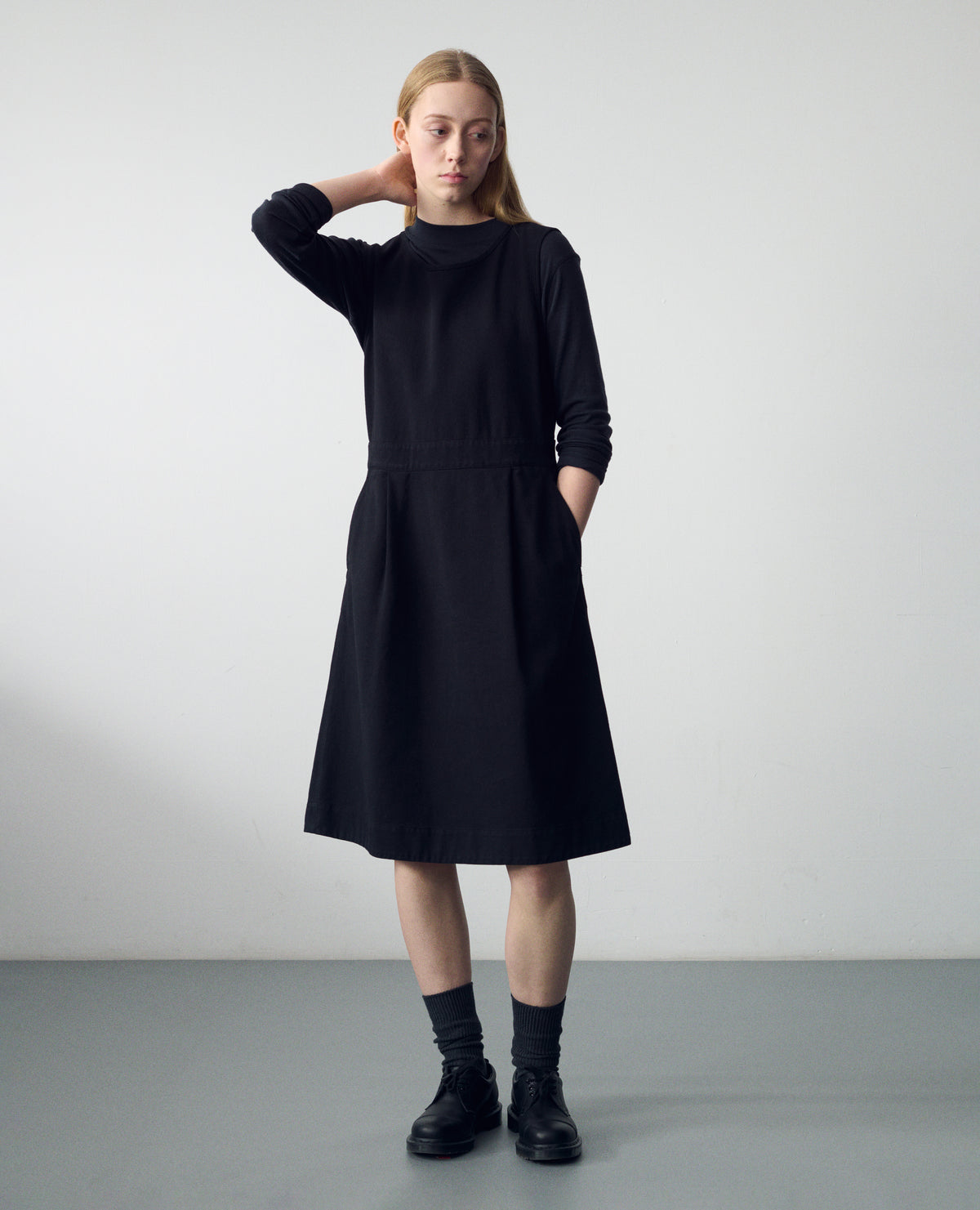 Full body image, front view of model wearing black sleeveless cotton dress, with side button fastening, two pleats and slanted pockets. Styled over a black long sleeve jersey top, black socks and leather shoes.