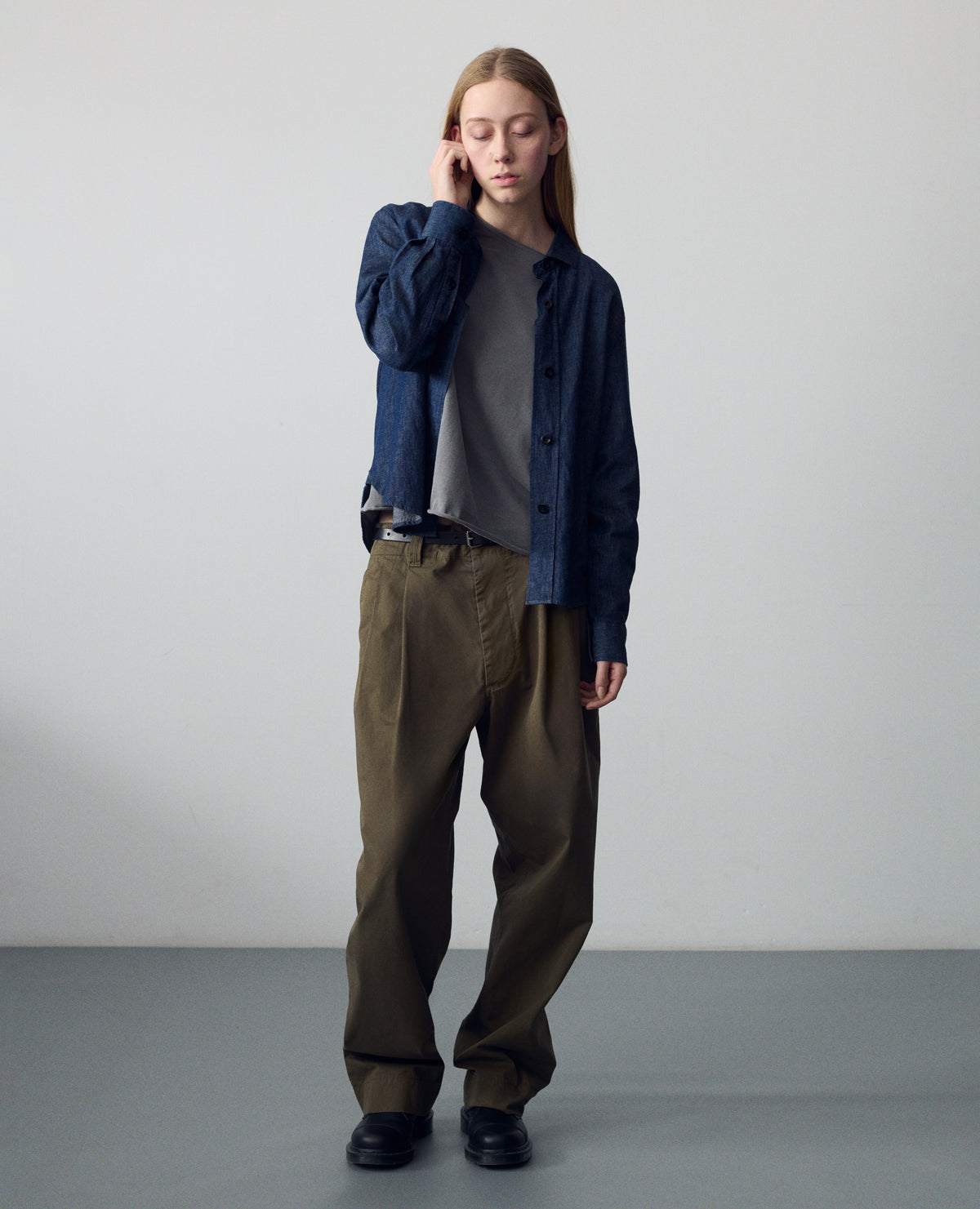 Full body image, front view of model wearing a dark blue shirt with a stitch-through raised placket, and a button-through cuff gauntlet. Styled with a grey jersey top, khaki green pleated trousers, black leather belt and leather shoes.