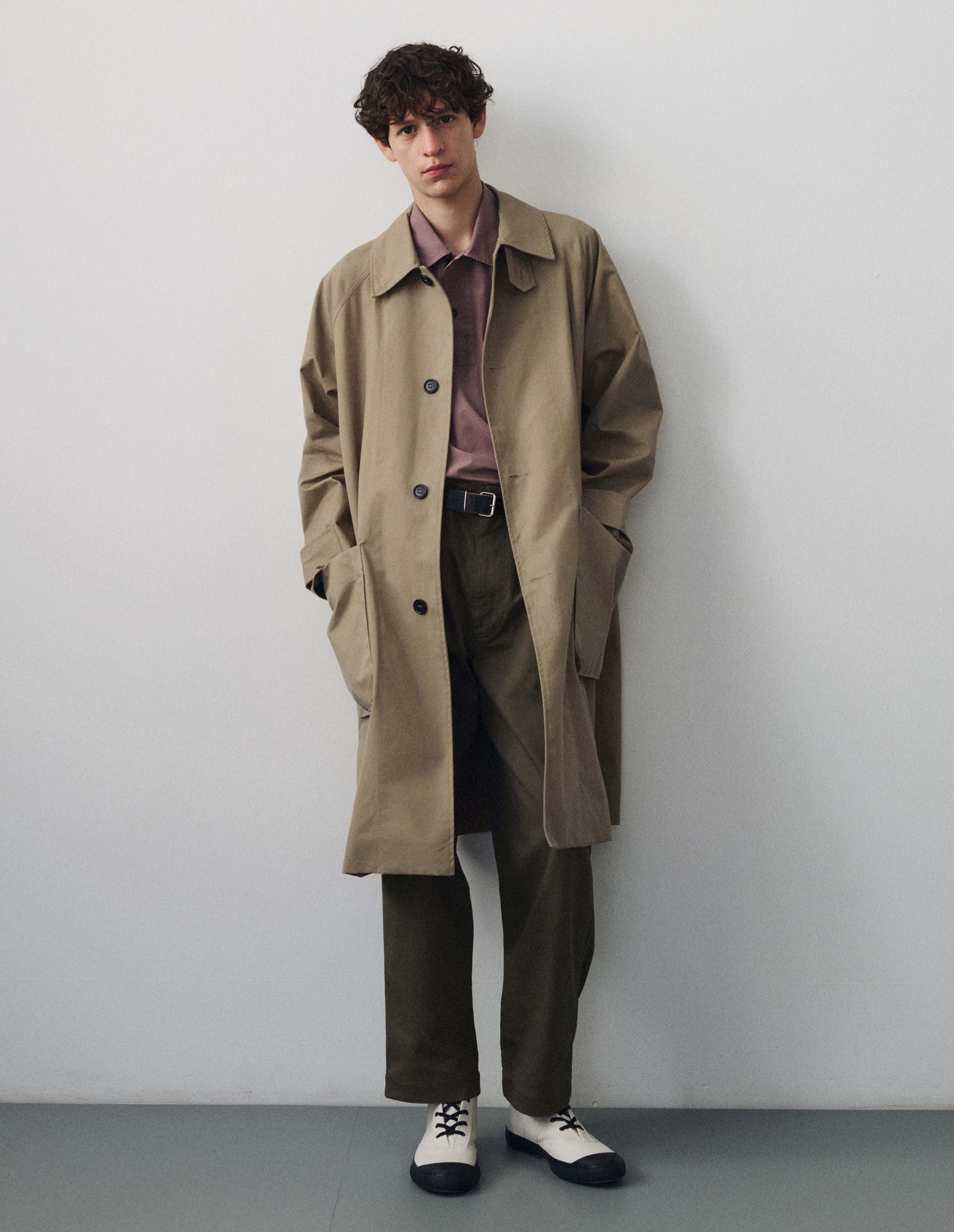 Full body image, front view of model wearing beige long sleeve cotton mac, with storm tab fastening neck, raglan sleeve and large patch bellow pockets. Styled with pink collared shirt, brownish green trousers and cream canvas trainers.