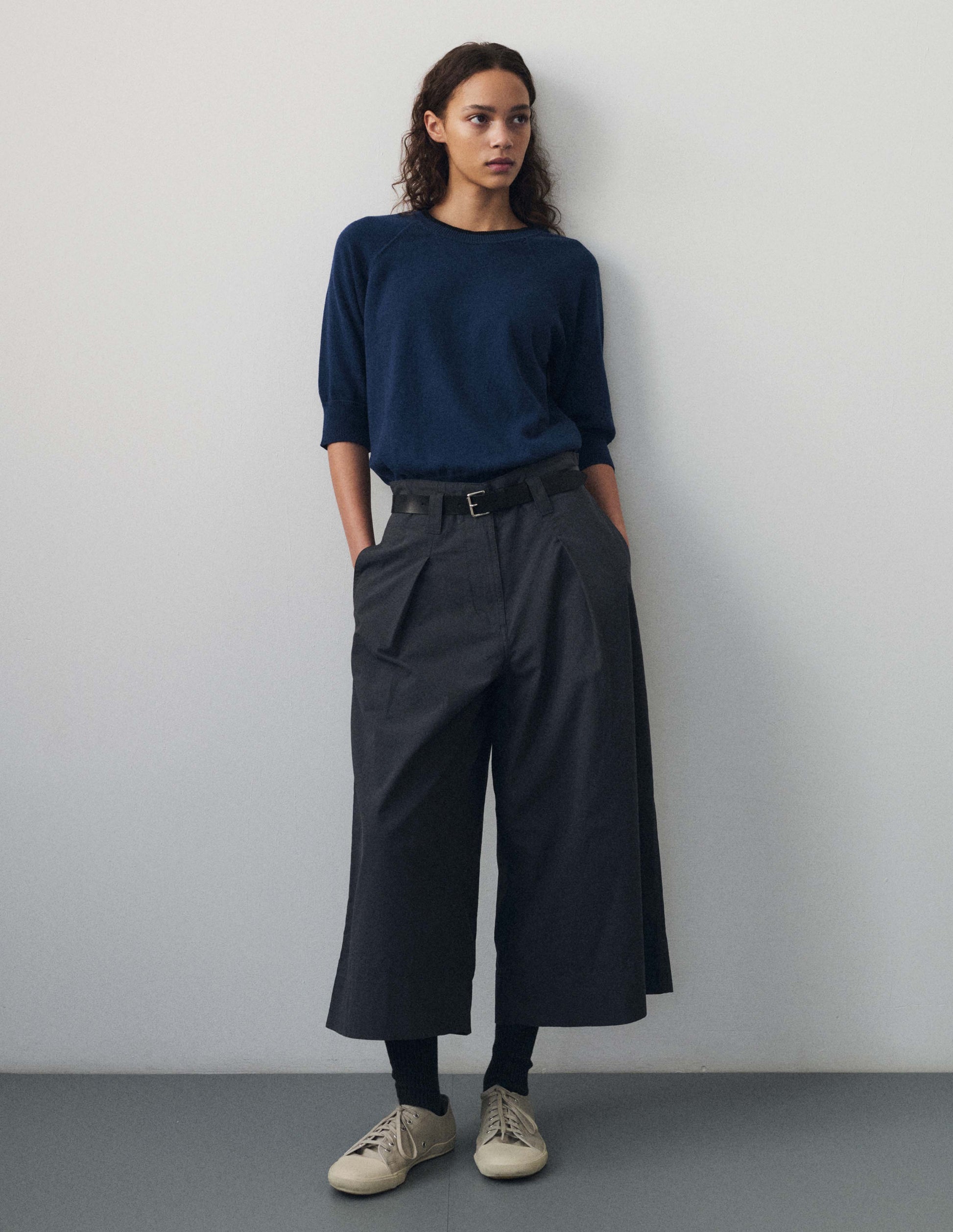 Full body image, front view of model wearing blue short sleeve knitted t shirt, with contrast black stitching on neckline, raglan sleeve, flatlock stitch seam detailing and contrast rib on sleeve and hem. Styled with dark navy blue trousers, black leather belt, black socks and beige canvas trainers. 