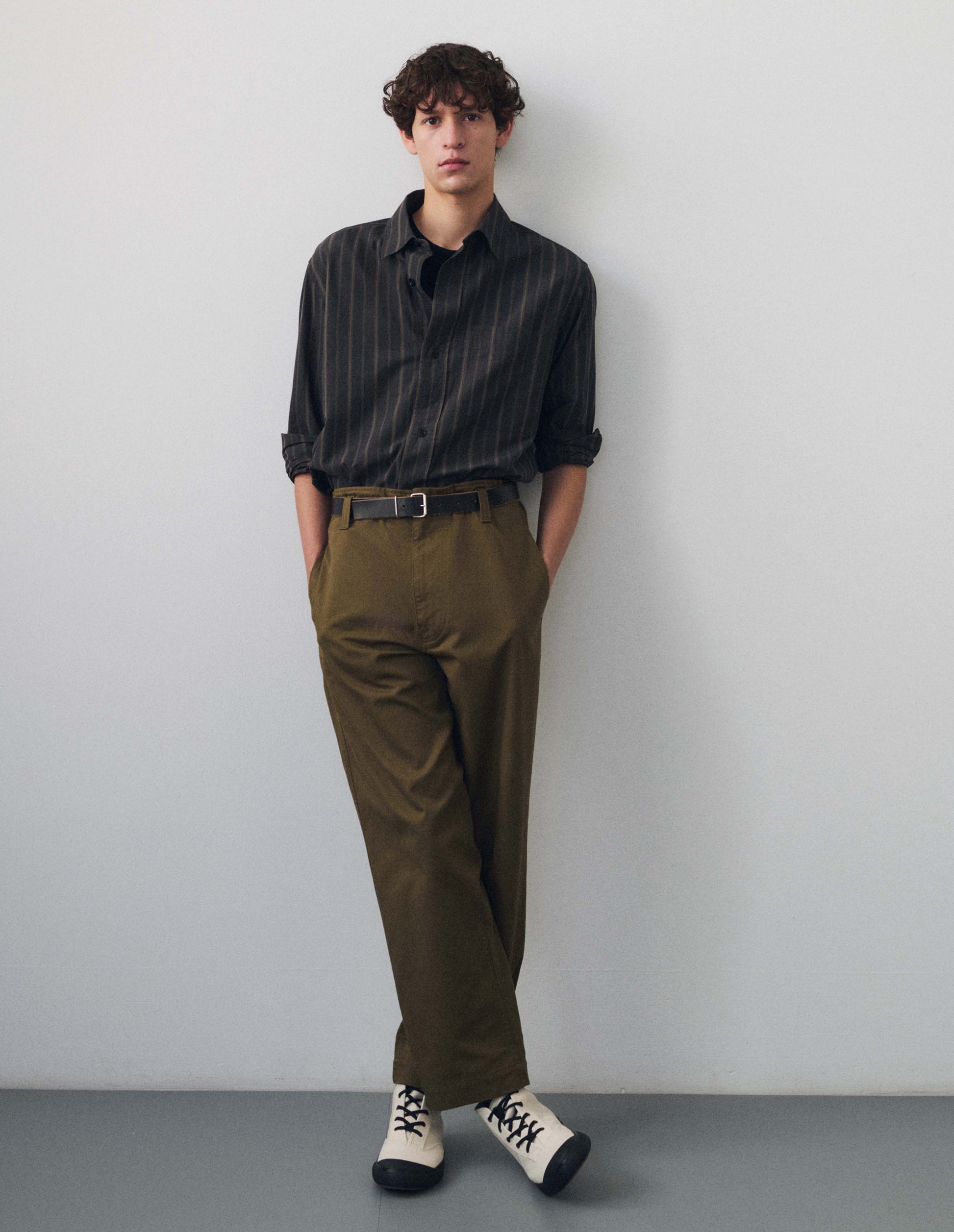 Full body image, front view of model wearing off black and khaki striped shirt, with corozo button fastening, patch chest pocket, contrast hanger loop on inside collar and elongated curved back hem. Styled with greenish brown trousers, dark brown leather belt and white canvas shoes with black soles.