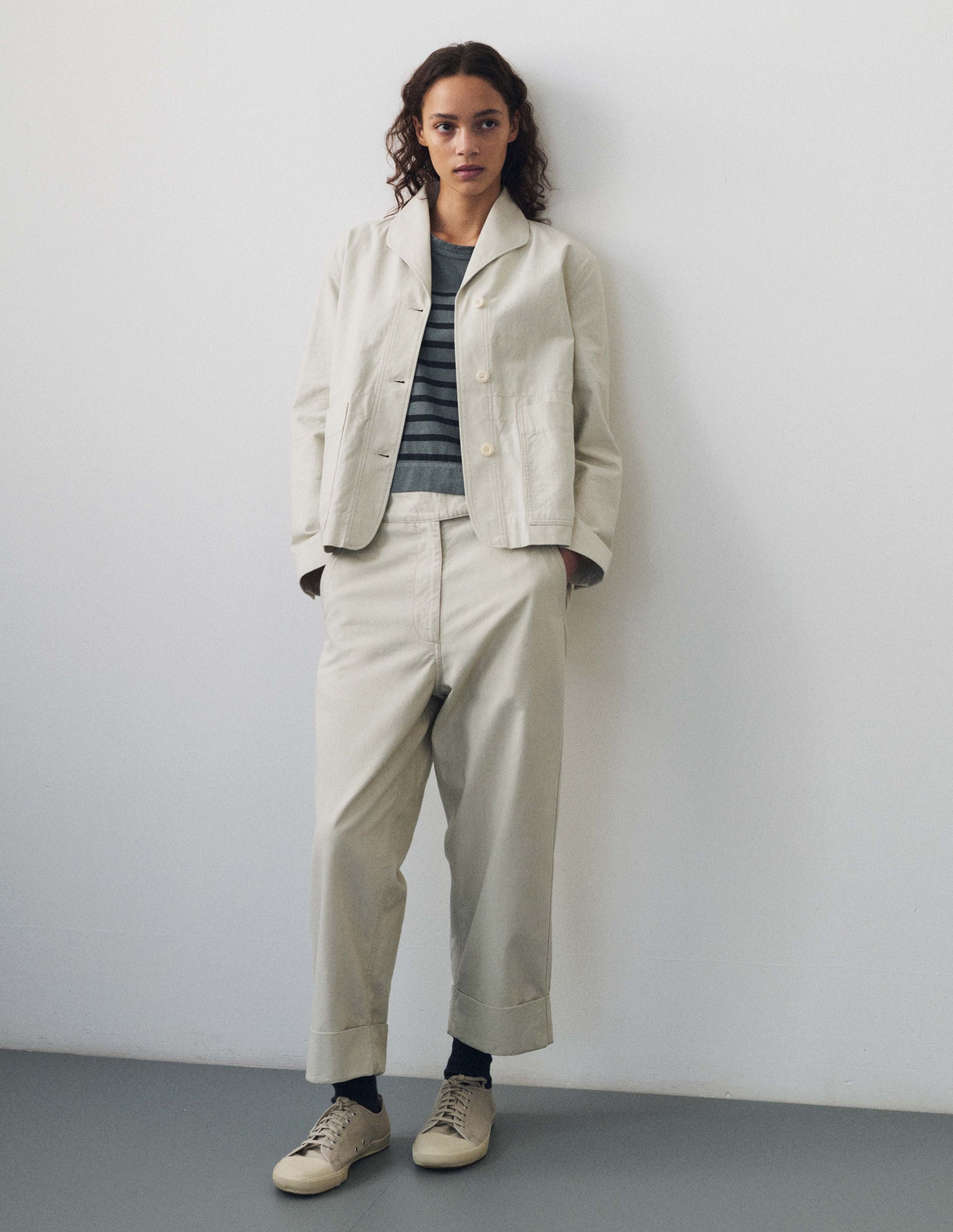 Full body image, front view of model wearing beige cotton drill trousers with slant pockets, corozo button detailing on waistband and turn up hems. Styled with matching beige blazer, blue and dark navy blue striped t shirt, black socks and white canvas trainers.