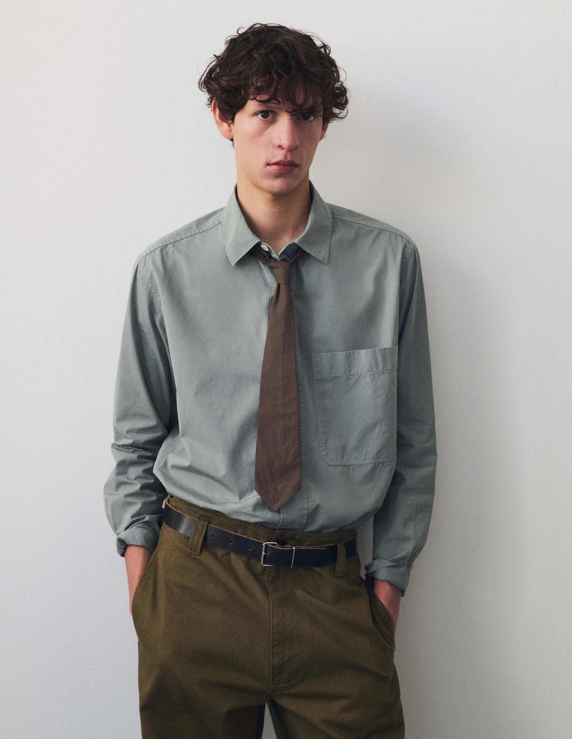 Full body image, front view of model wearing grey shirt, raised placket, pale corozo button fastening, large chest pocket, side gusset stitch detailing and hanger loop detailing on inside collar. Styled with greenish brown trousers, greyish brown tie and dark brown leather belt. 