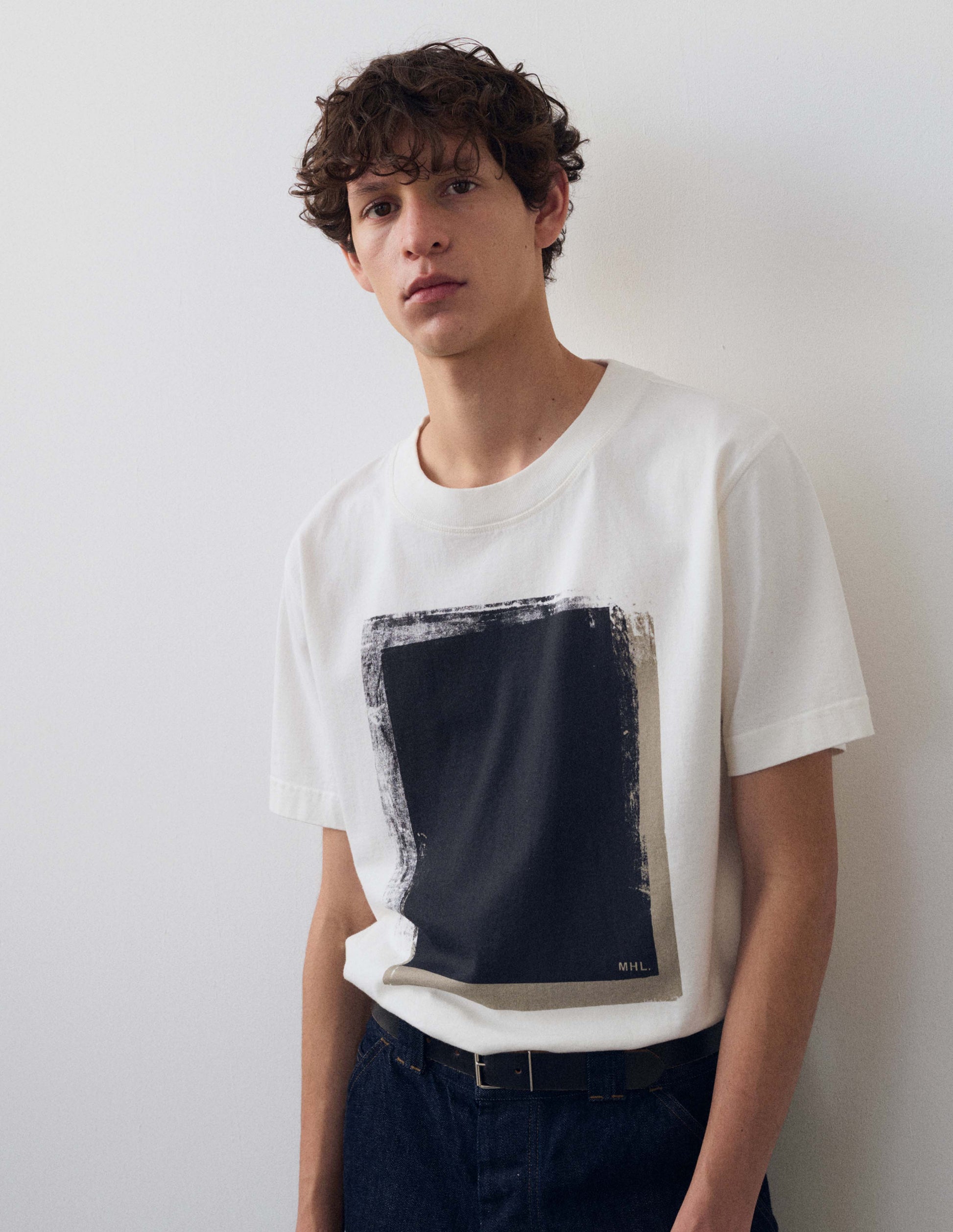 Full body image, front view of model wearing off white short sleeve t shirt with black abstract block printed on the front. Styled with dark blue jeans and dark brown leather belt. 