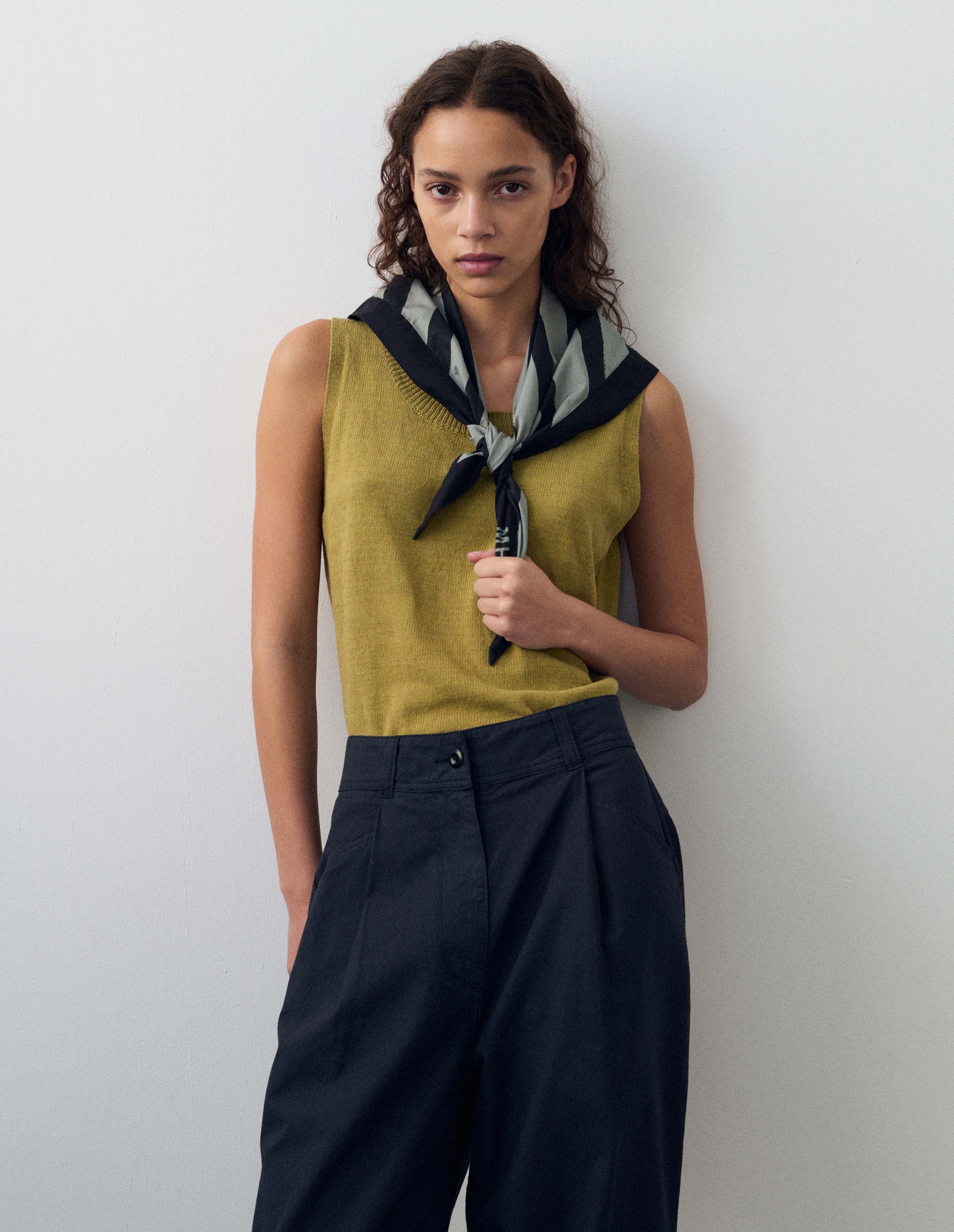 Cropped body image, front view of model wearing yellowish green knitted tank top with single rib crew neck and. Styled with dark navy blue trousers and pale blue neckerchief with navy stripes.    